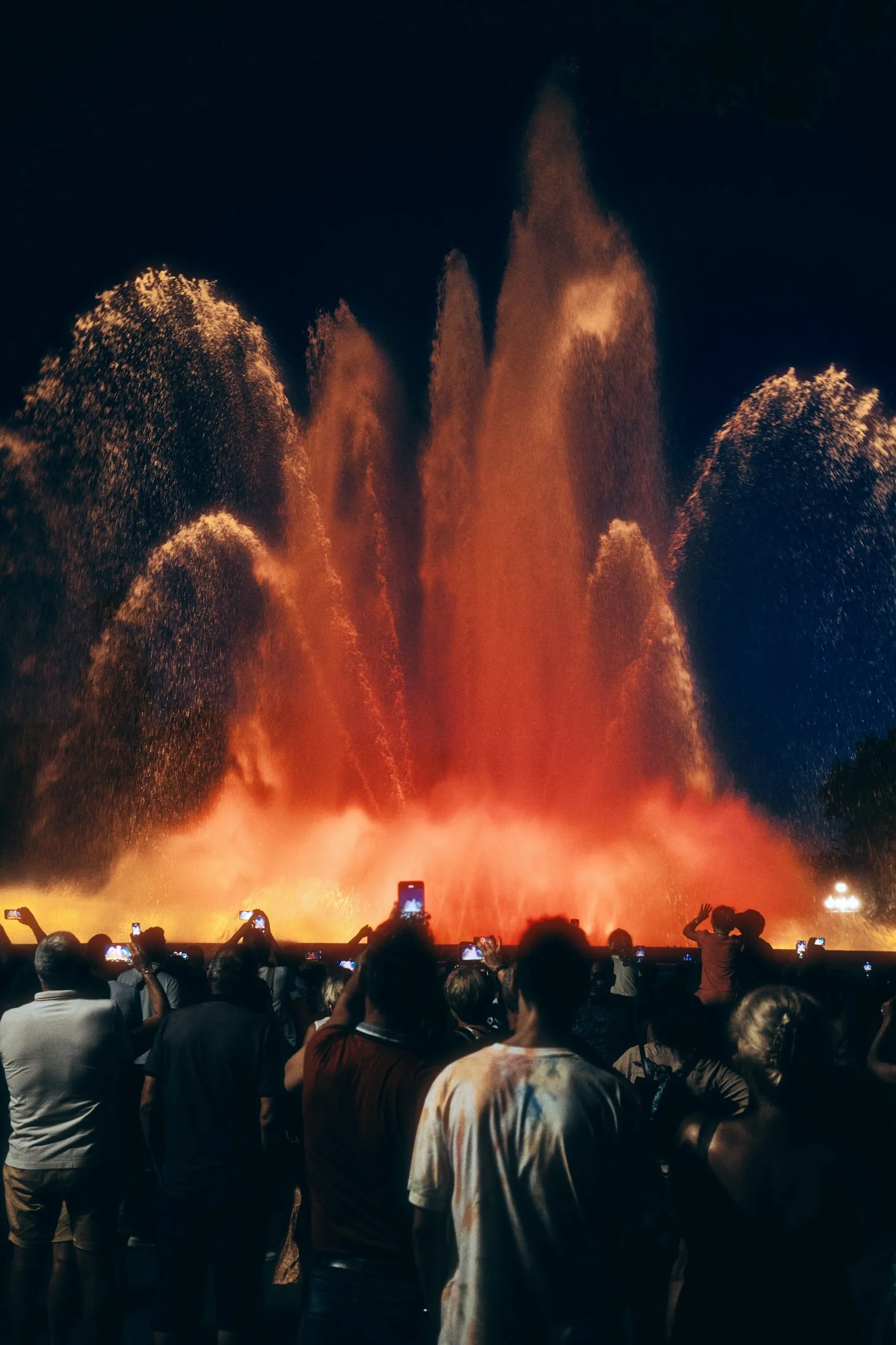 Travel photograph of the Magic Fountain of Montjuïc in Barcelona, Spain, illuminated at night with vivid red and orange water displays, framed by silhouettes of a crowd capturing the moment on their phones. Captured by Miami and Los Angeles-based com