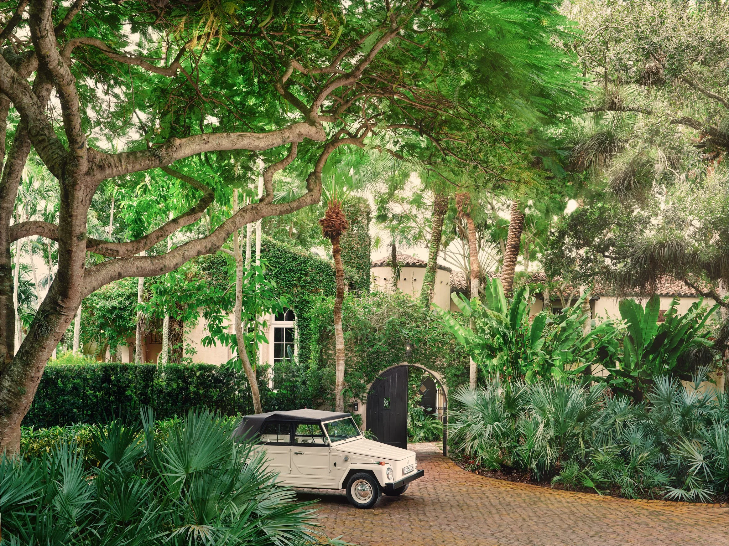 Architectural photograph of Villa Serenita's lush tropical driveway in Miami, featuring a vintage white Volkswagen Thing parked on a brick drive under the canopy of mature trees, with the home's Mediterranean Revival facade partially visible. Capture