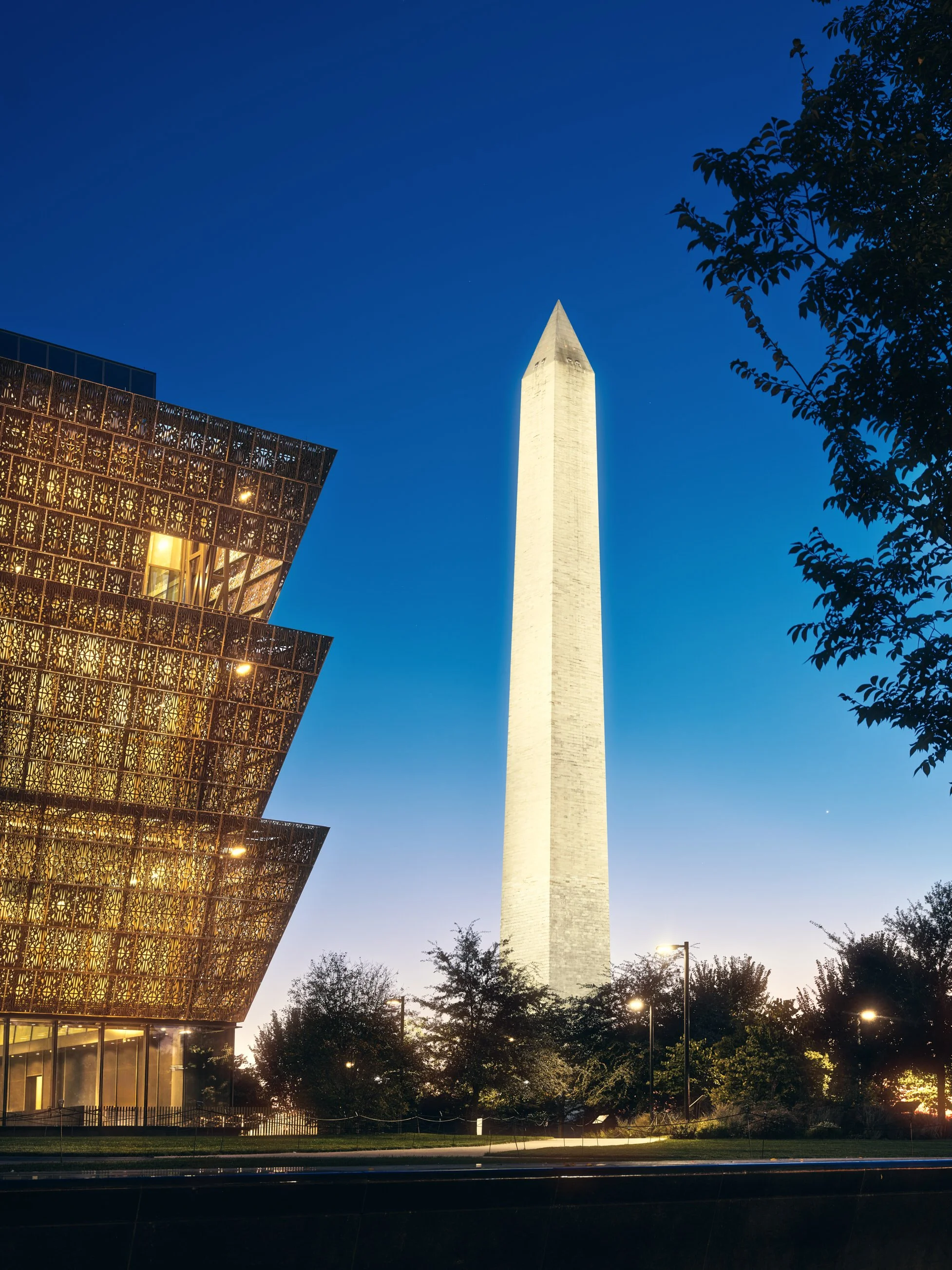 Architectural photograph of the Smithsonian National Museum of African American History and Culture in Washington, D.C., taken at twilight. The museum’s bronze lattice façade glows against a vibrant blue and purple sky, with the illuminated Washingto