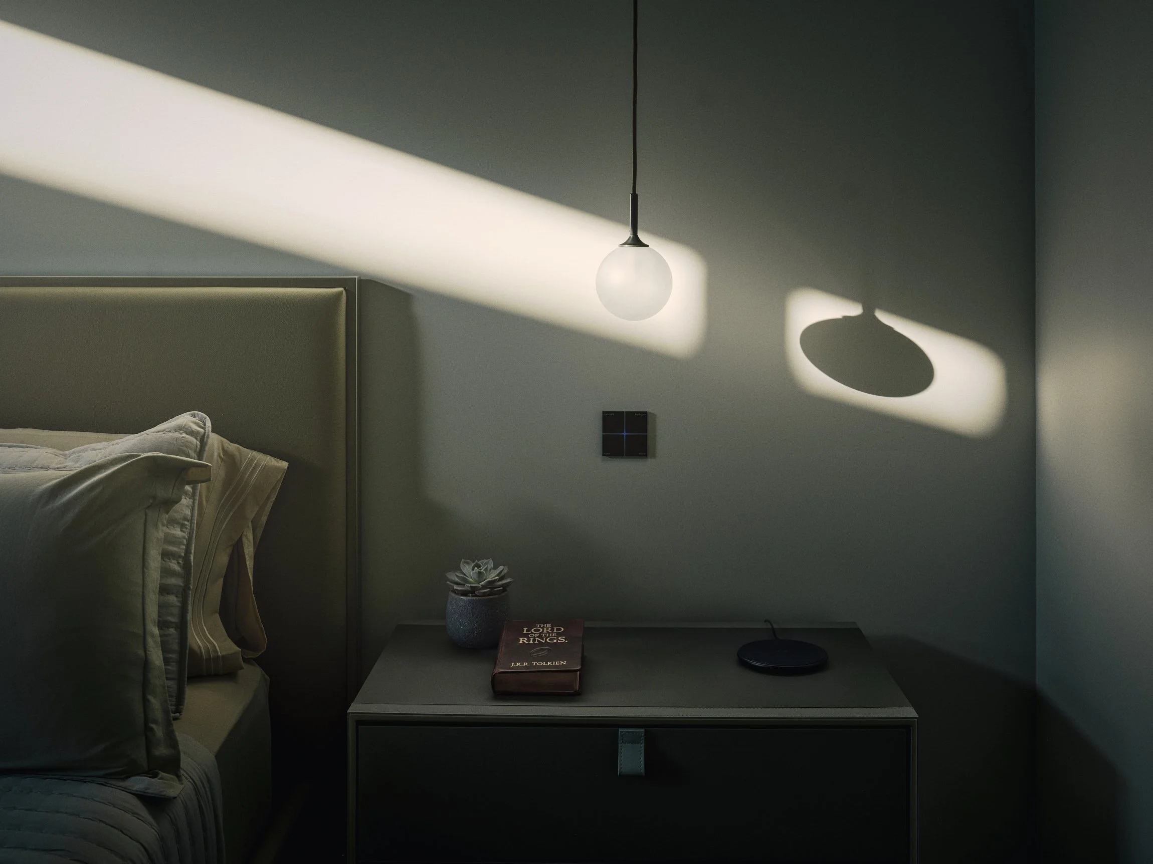 Architectural photograph of a bedroom light fixture and night stand. The light is moody with a single streak of sunlight cutting through the wall in a dramatic pattern and marking the translucent orb-shaped bulb. Taken by Miami architectural photogra