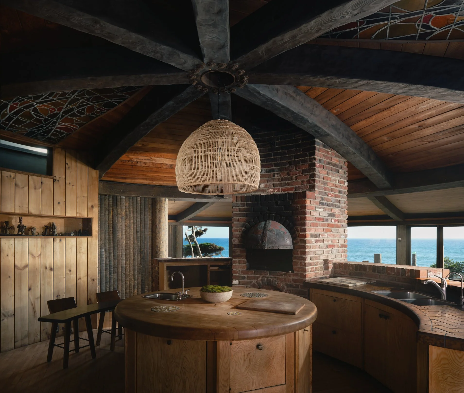 Architectural photograph of the Sandcastle House kitchen in Malibu, designed by architect Harry Gesner. The round, wood-paneled kitchen features custom curved cabinetry, a central circular island, a brick pizza oven, and exposed wood beams fanning ac