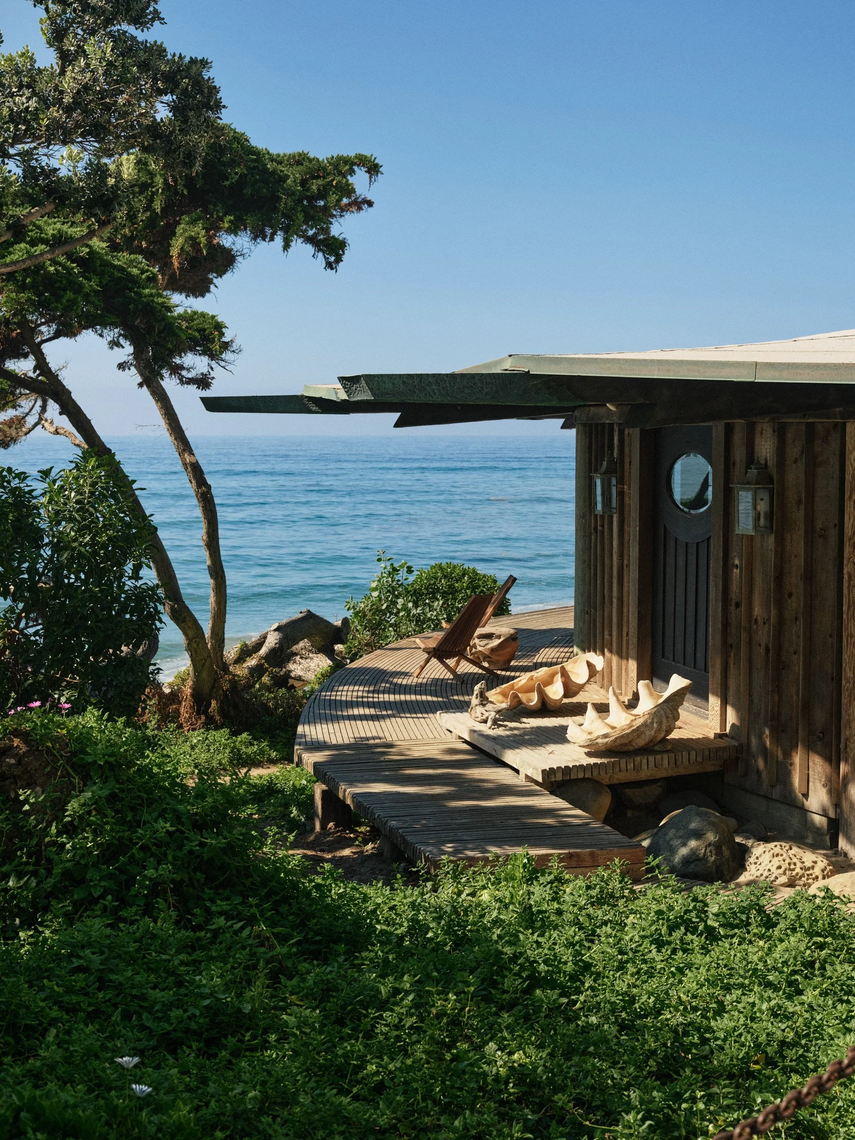 Architectural photograph of the Sandcastle House in Malibu, California, showcasing its curving wooden boardwalk, oceanfront setting, and natural materials like reclaimed telephone poles and driftwood. Designed by architect Harry Gesner for his wife,