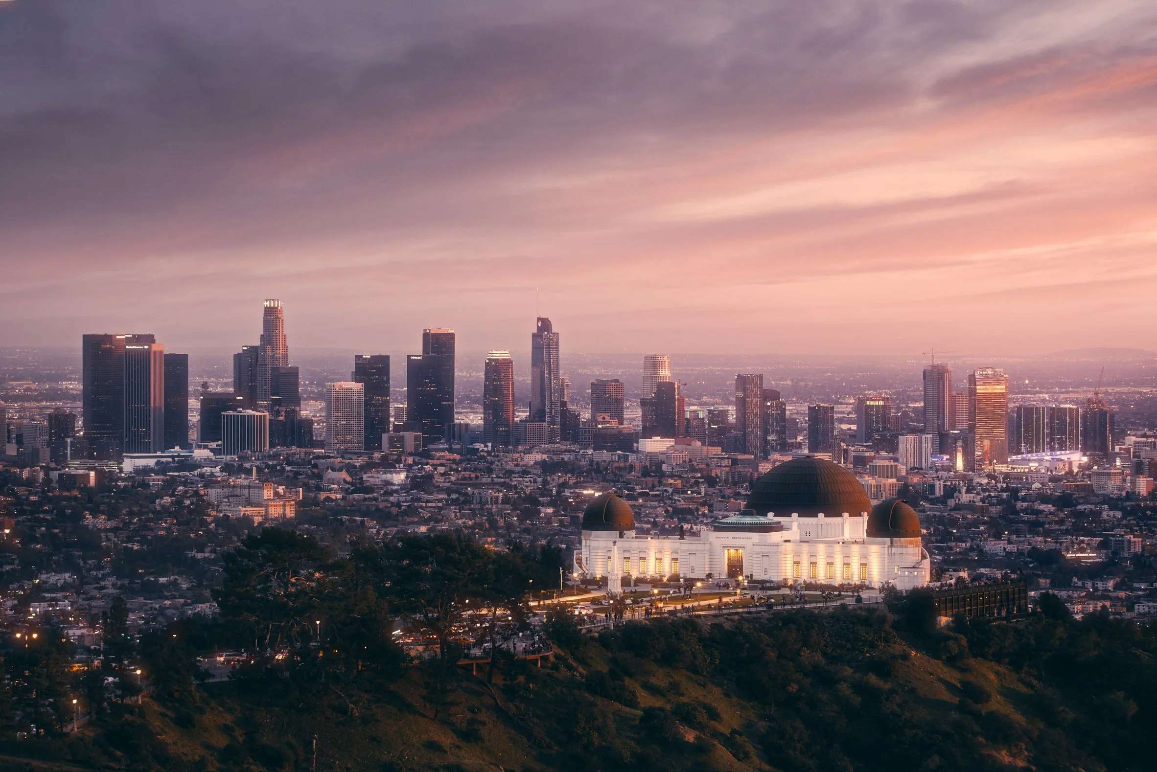 Architectural travel photograph of Griffith Observatory illuminated at twilight with the Los Angeles skyline in the background, captured under a soft pink and purple sunset sky. The scene highlights the contrast between the historic landmark in the f