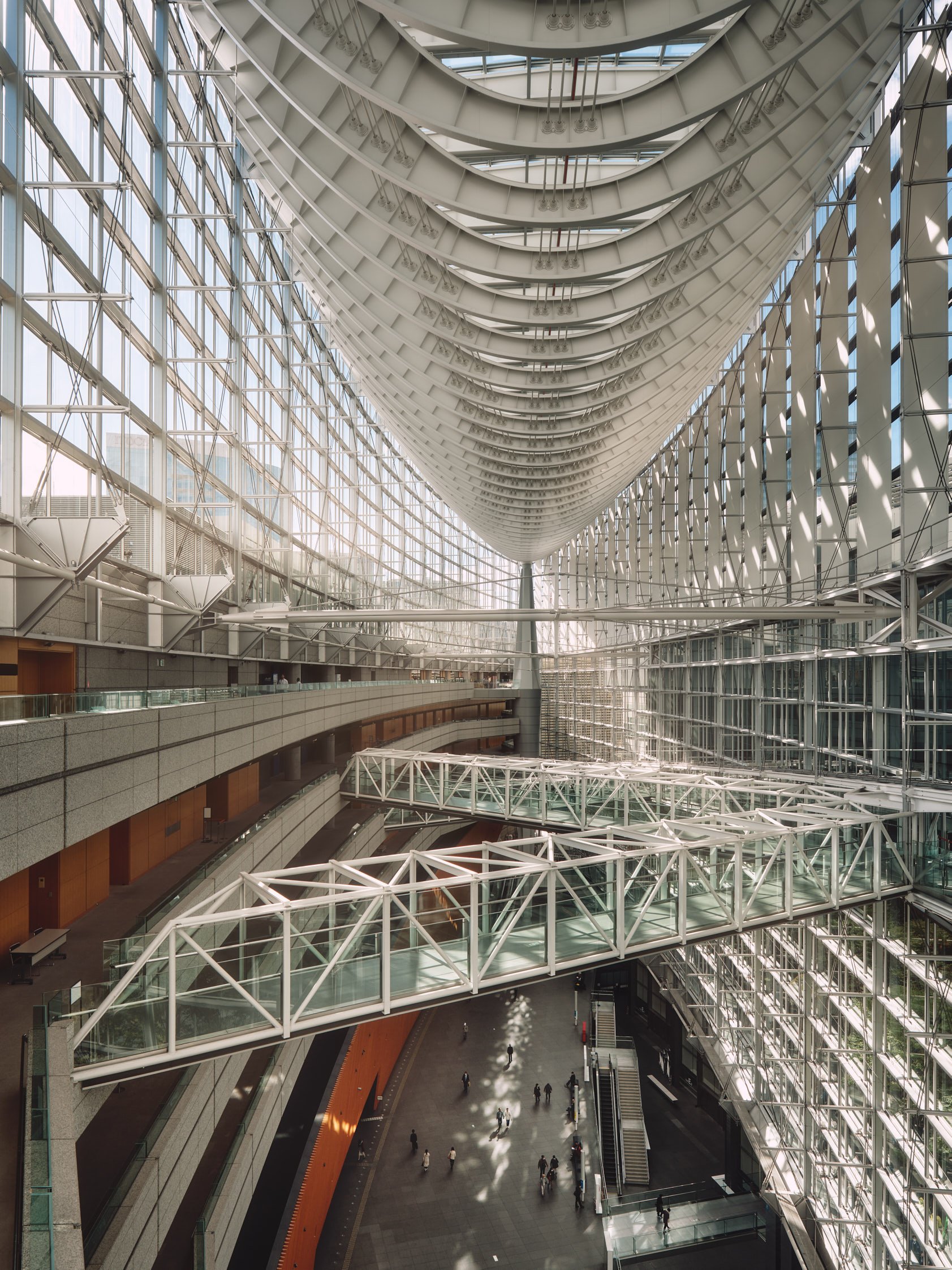 Architectural photograph of the Tokyo International Forum in Japan, highlighting its iconic glass atrium and sweeping steel structure. The image captures the dramatic symmetry and scale of the interior, with pedestrian bridges suspended above the con