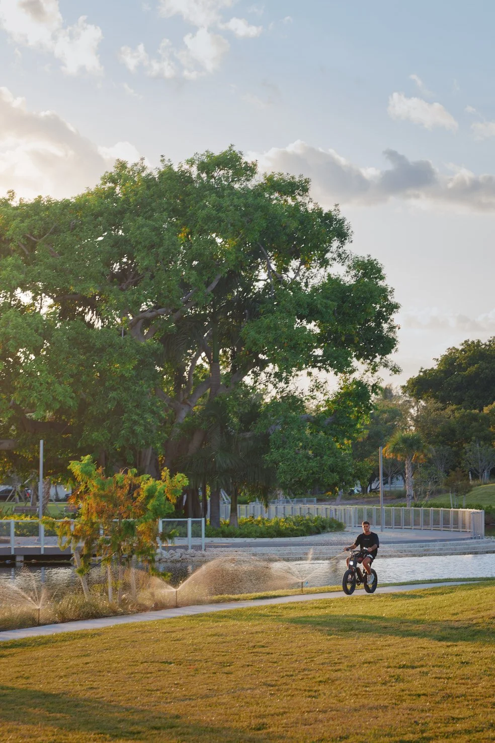 Architectural photograph of a cyclist riding along a waterfront path beside the lake in Bayshore Park in Miami Beach, with irrigation sprinklers and restored landscape framing the park’s pedestrian and bicycle circulation network. Captured by Miami a