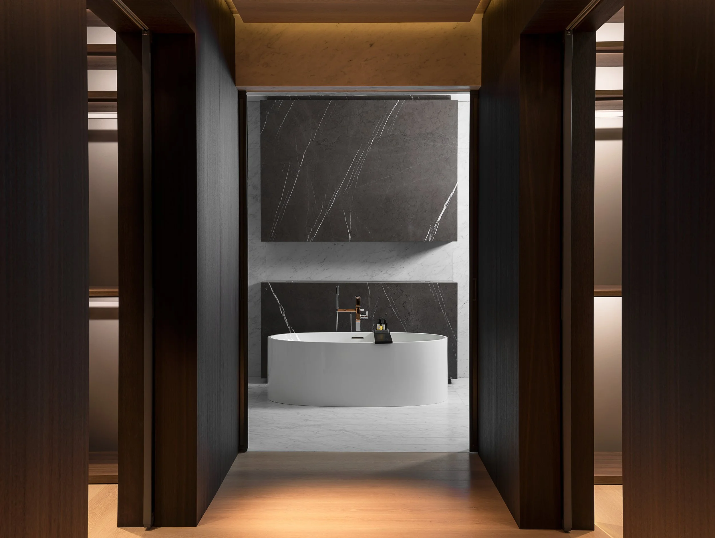 Architectural photograph of a luxury bathroom interior featuring a freestanding oval bathtub framed by dark wood paneling and illuminated walk-in closets, with large marble slabs in gray and white adding a dramatic backdrop. Captured by Miami and Los
