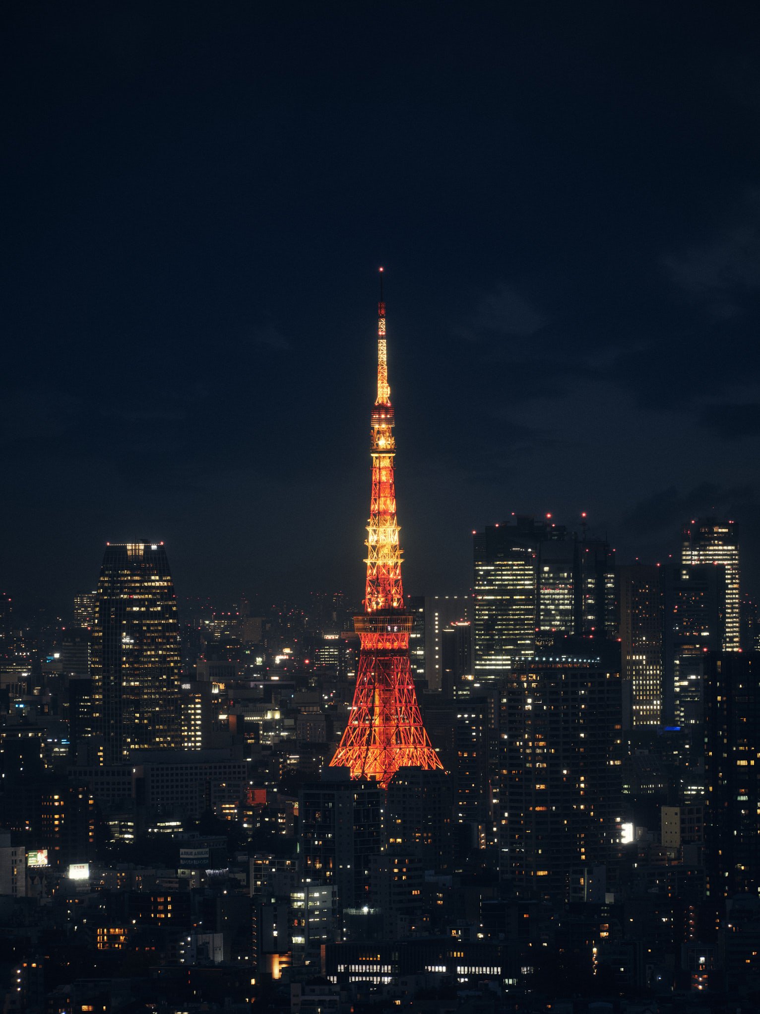 Architectural photograph of Tokyo Tower illuminated in vibrant red and orange hues at night, rising above the city skyline in Minato, Tokyo. Surrounded by high-rise buildings and a glowing urban grid, the iconic communications and observation tower s