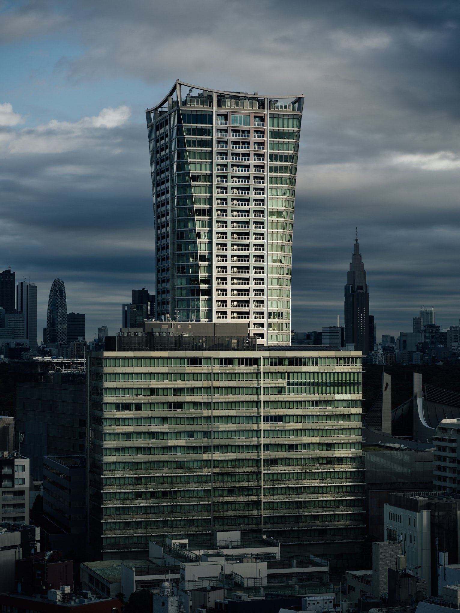 Architectural photograph of a striking modern tower in Tokyo featuring a flared rooftop and glass facade, captured under dramatic overcast skies. The city’s layered skyline—including Mode Gakuen Cocoon Tower and the NTT Docomo building—adds urban dep