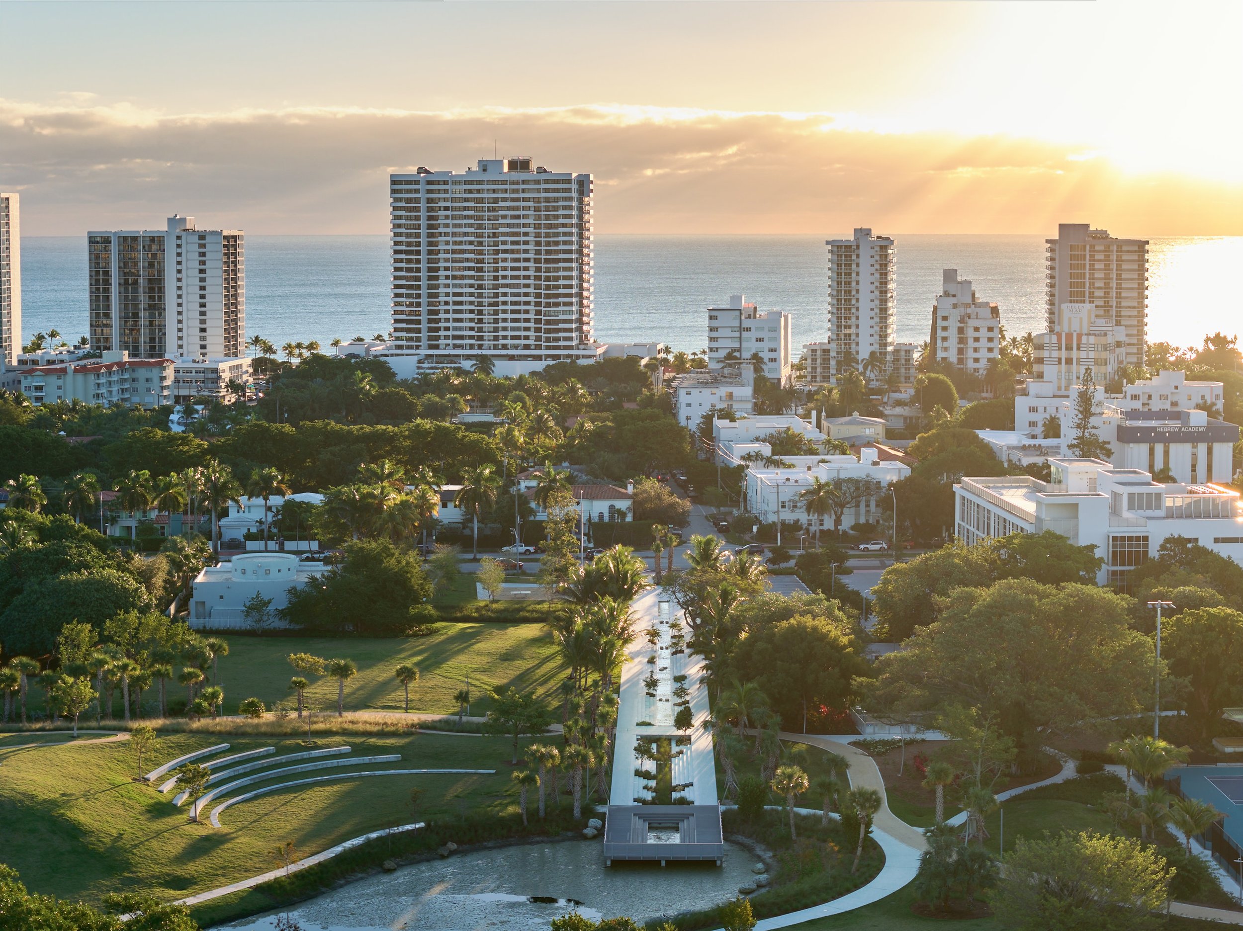 Architectural aerial photograph of Bayshore Park in Miami Beach with the Atlantic Ocean beyond, highlighting the park’s amphitheater lawn, pedestrian bridge, and surrounding urban landscape at Sunrise. Captured by Miami and Los Angeles-based architec