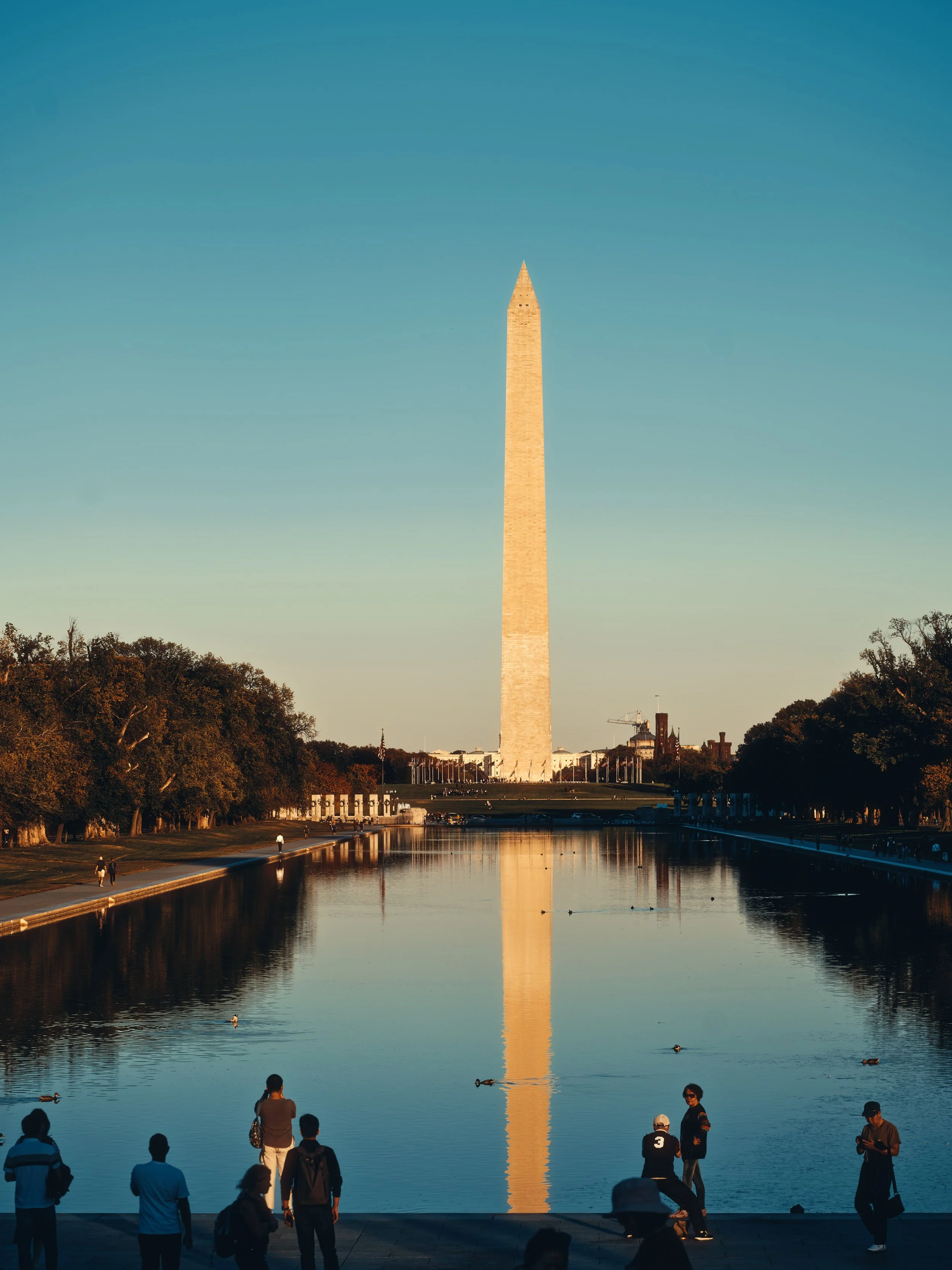 Architectural, travel photograph of the Washington Monument reflecting in the Lincoln Memorial Reflecting Pool at golden hour in Washington, D.C., with a clear blue sky and warm sunlight creating a serene, symmetrical composition. Captured by Miami a