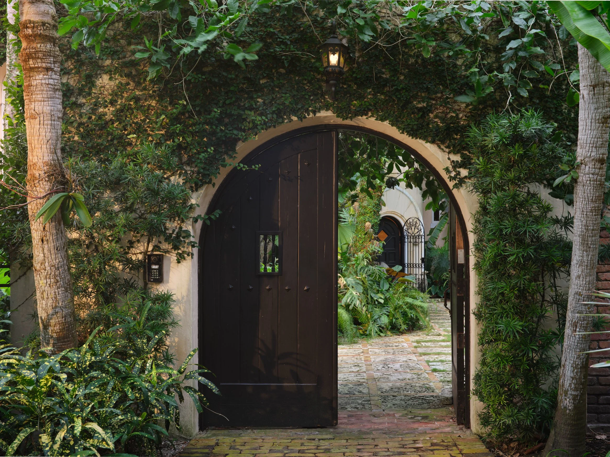 Architectural photograph of a Mediterranean-style arched wooden gate partially open, framed by lush tropical foliage and climbing ivy, revealing a stone pathway leading into a verdant courtyard. Captured by Miami & Los Angeles architectural & travel