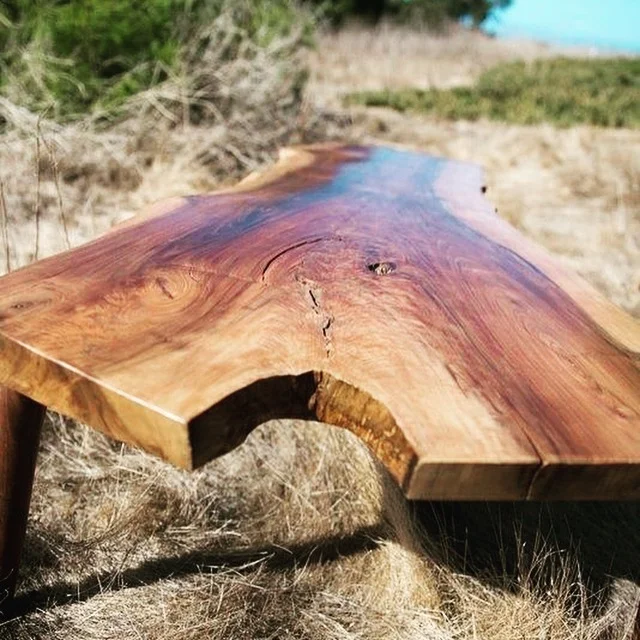 Close-up of a polished wooden outdoor table showing wood grain and natural edge, with grass and sandy shore in the background near the water.