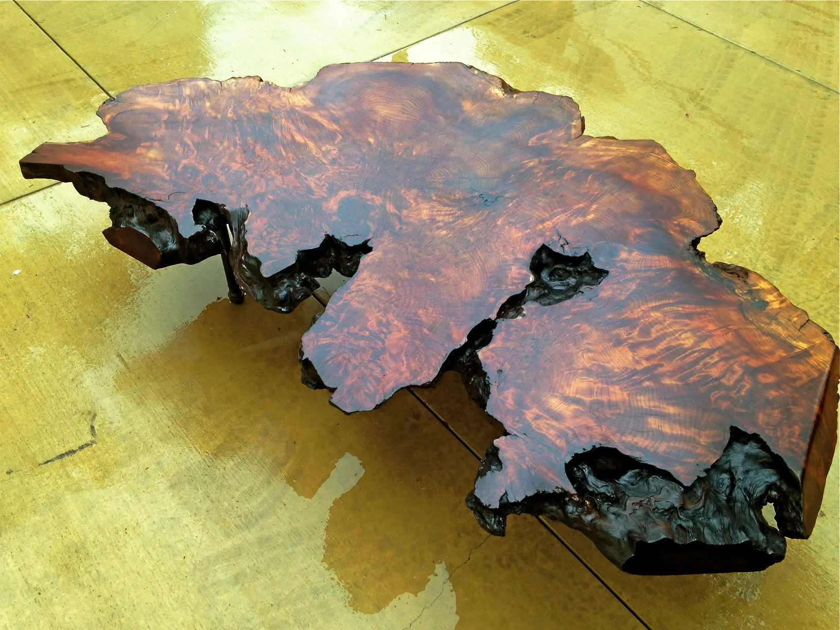 A wooden table with a natural, irregular shape showing a rich, reddish-brown grain and a shiny finish, placed on a yellowish tiled floor.