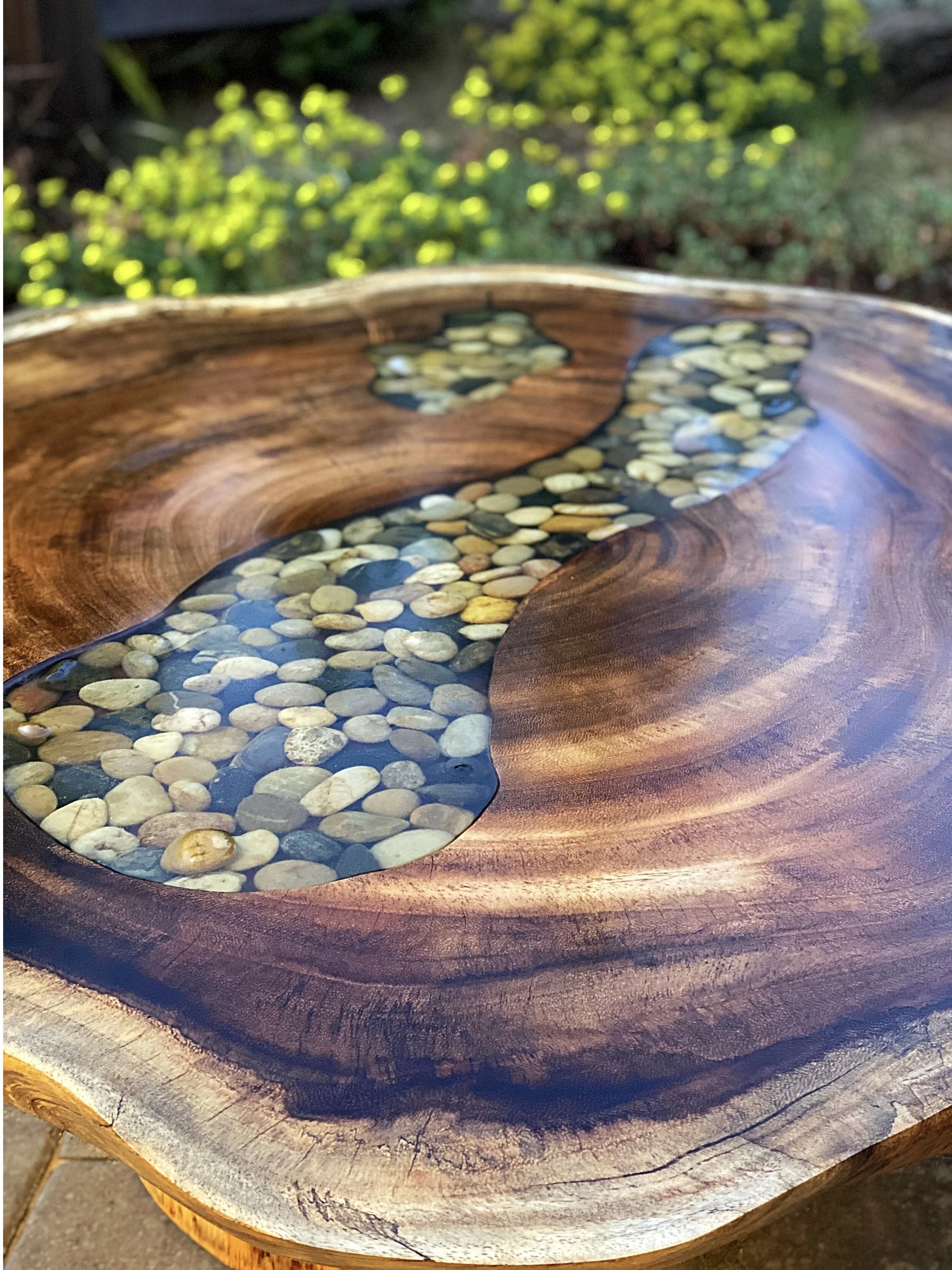 Close-up of a wooden table with a natural edge, featuring a carved river design filled with small, rounded pebbles and water.