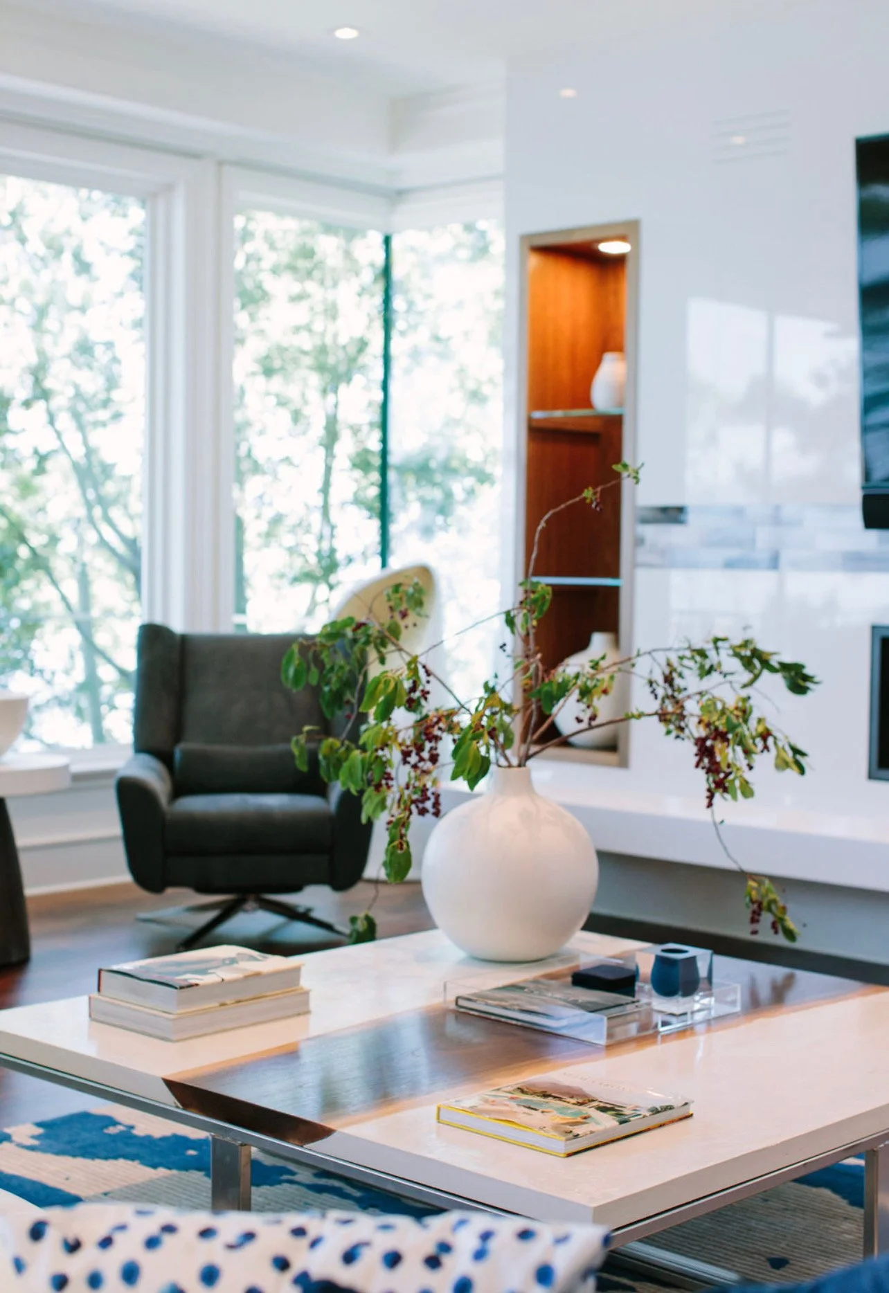 Living room with a white coffee table, a white vase with green and red-stemmed branches, an armchair, a built-in shelf, large windows with trees outside, and modern decor.