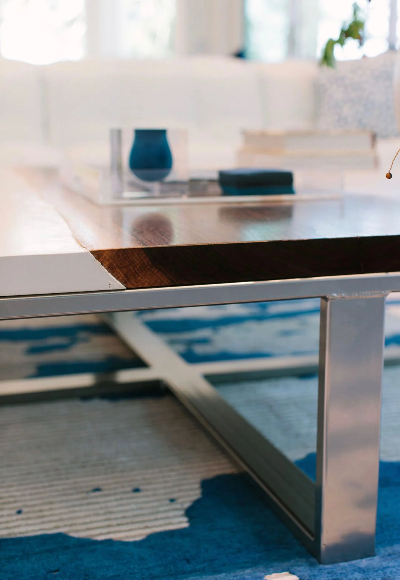 Close-up of a modern table with a wooden top and metallic legs, placed on a patterned rug in a bright, well-lit room.
