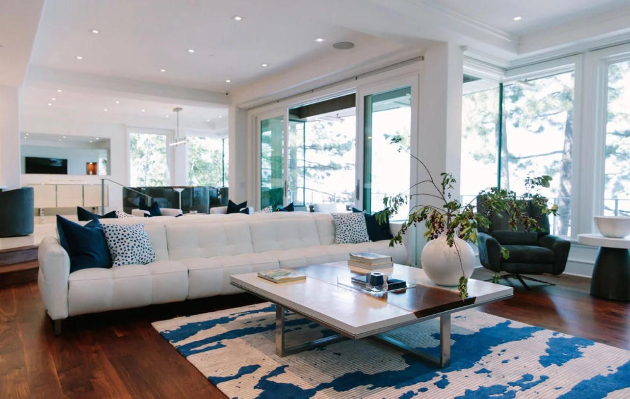 Modern living room with white sofa, black and polka dot pillows, wooden coffee table with books and a white vase with green foliage, black armchair, large windows with trees outside, hardwood floors, and recessed ceiling lights.
