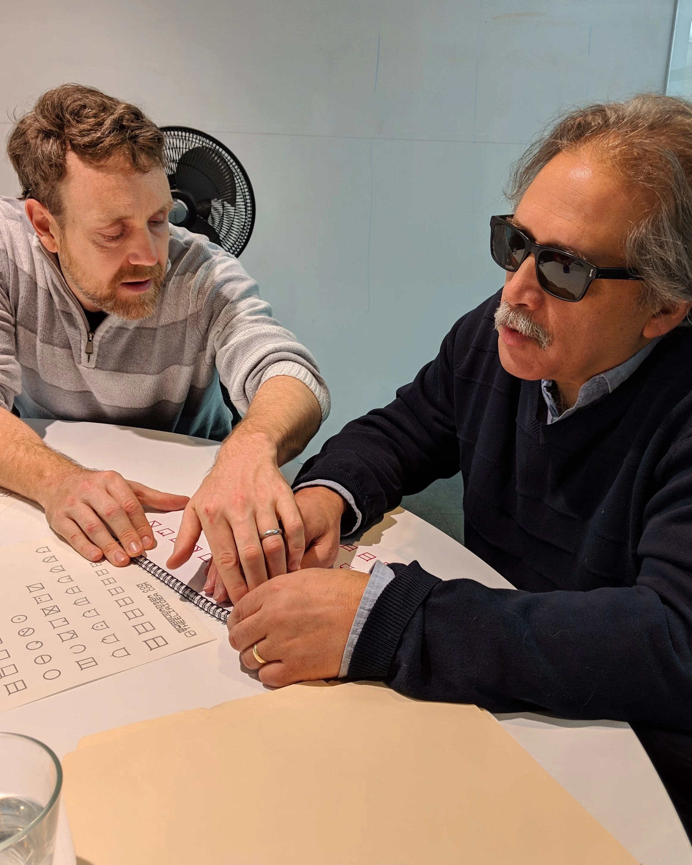 Adam (on the left) using his hands to show Tom (on the right) the letter “C”. Tom and Adam are both previously sighted.