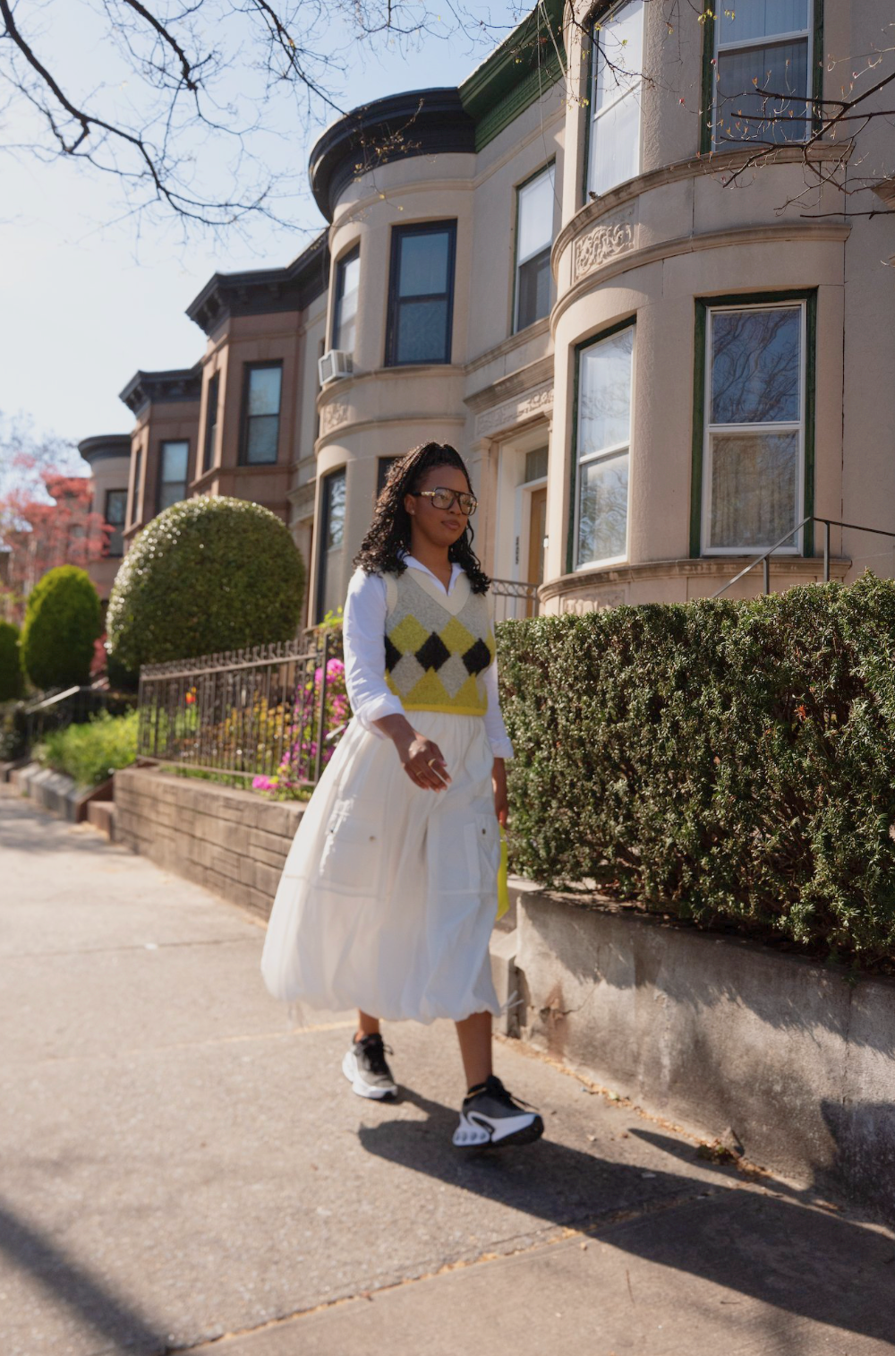 Woman walking on sidewalk in a suburban neighborhood with historic brownstone houses, wearing glasses, a white skirt, a white shirt with a yellow and black argyle vest, and black and white sneakers.