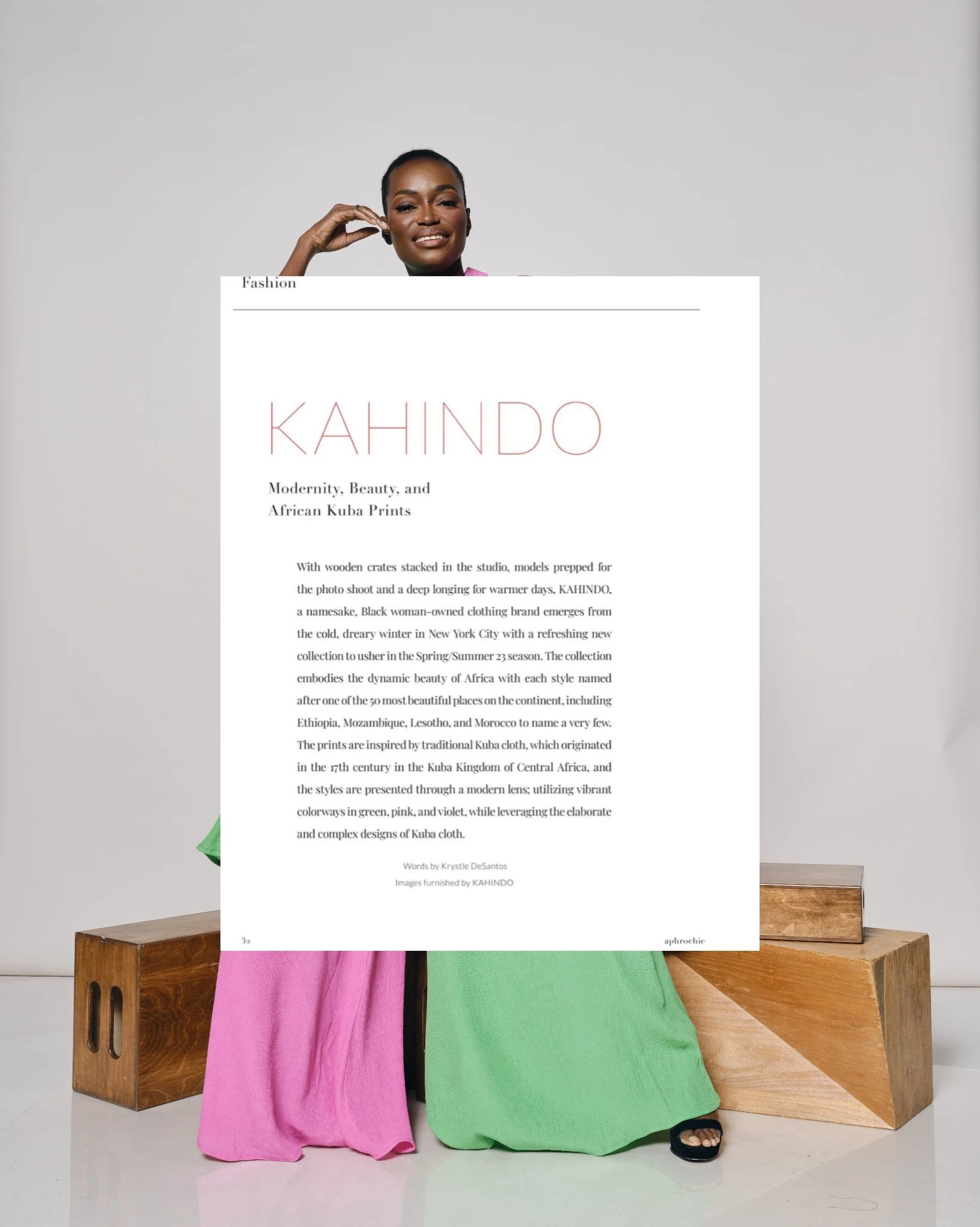 A woman standing behind a large white paper or poster that covers most of her face and body, with only her chin, eyes, and left hand visible. She is wearing a pink and green dress and standing next to wooden blocks. The poster features the word 'KAHINDO' in large pink letters and includes text about African Kuba prints and fashion.