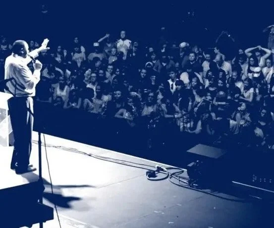 A man on stage speaking or singing to a large audience in a dimly lit venue.