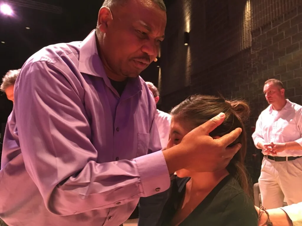 A man with a purple shirt gently holds a woman's head, preparing to perform a healing or prayer ritual on her forehead in a dimly lit indoor setting, with other people in the background.