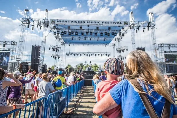 Crowd watching a live performance at the NightVision Music Festival, outdoor stage with scaffolding and sound equipment, partly cloudy sky
