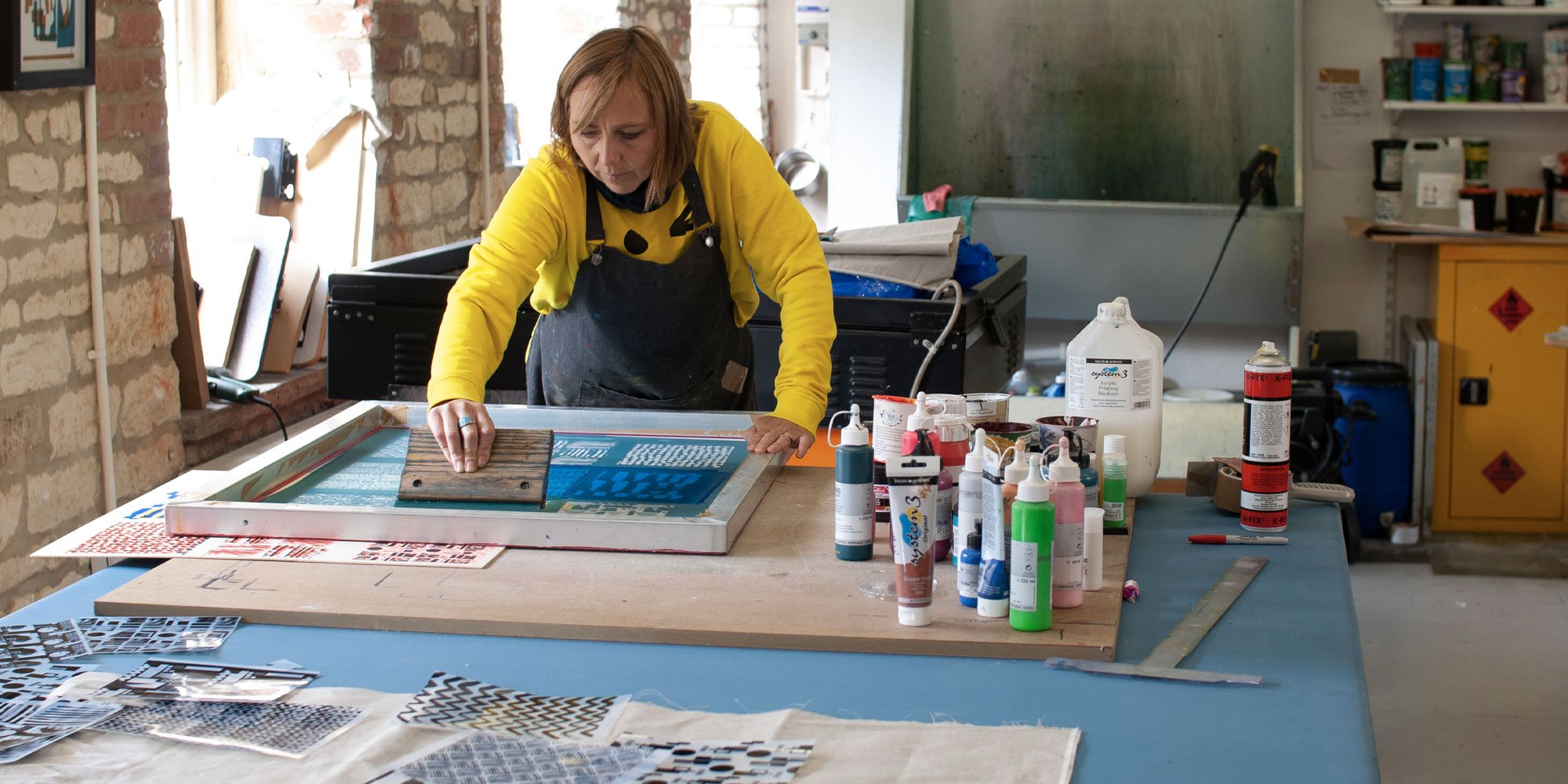 Woman working on a screen printing project at a large worktable in a studio with art supplies and tools around.