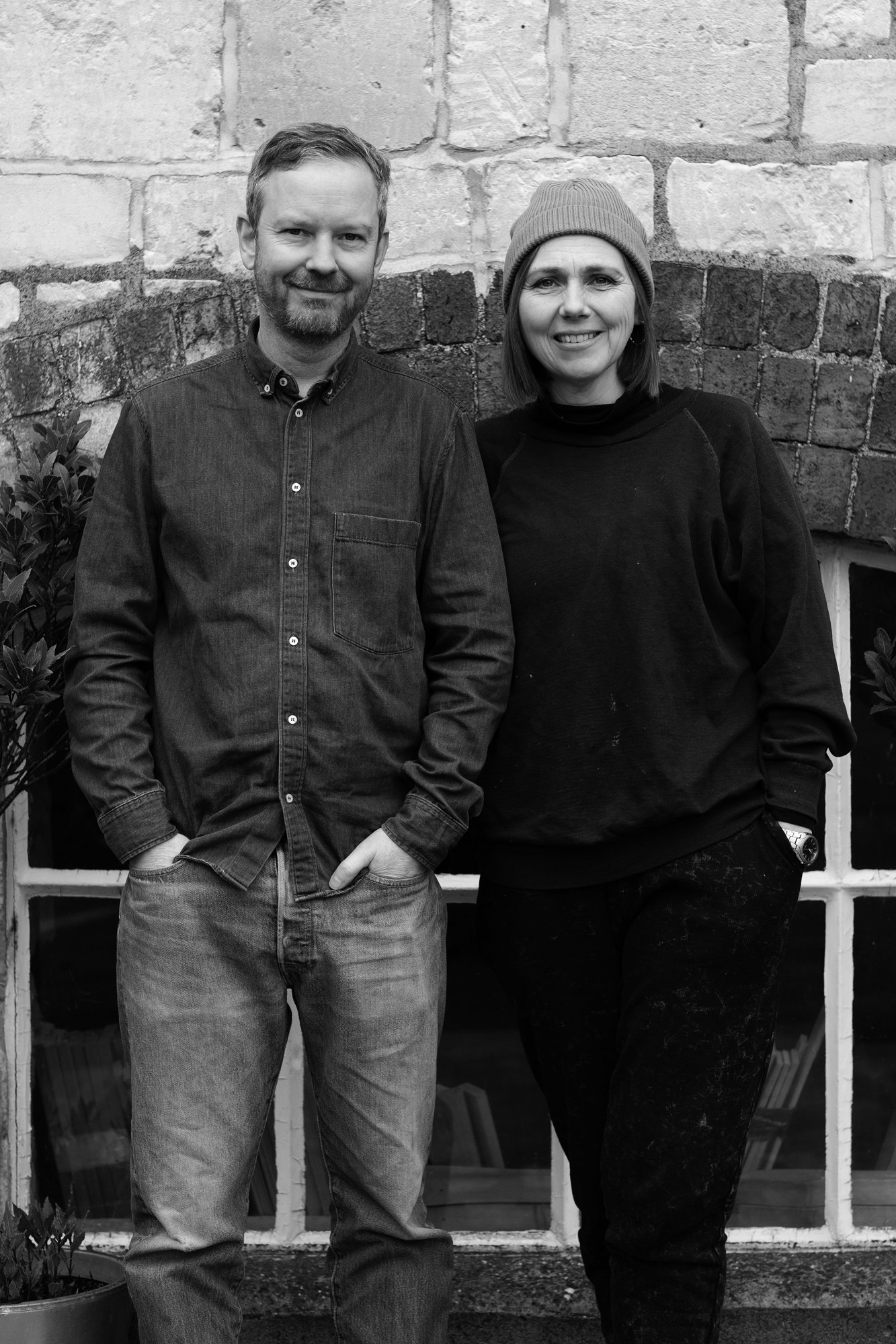 standing outdoors in front of a brick wall and window. The man is wearing a dark shirt and jeans, with his hands in his pockets. The woman is wearing a beanie, black sweatshirt, and dark pants, with her hands in her pockets. The photo is in black and white.