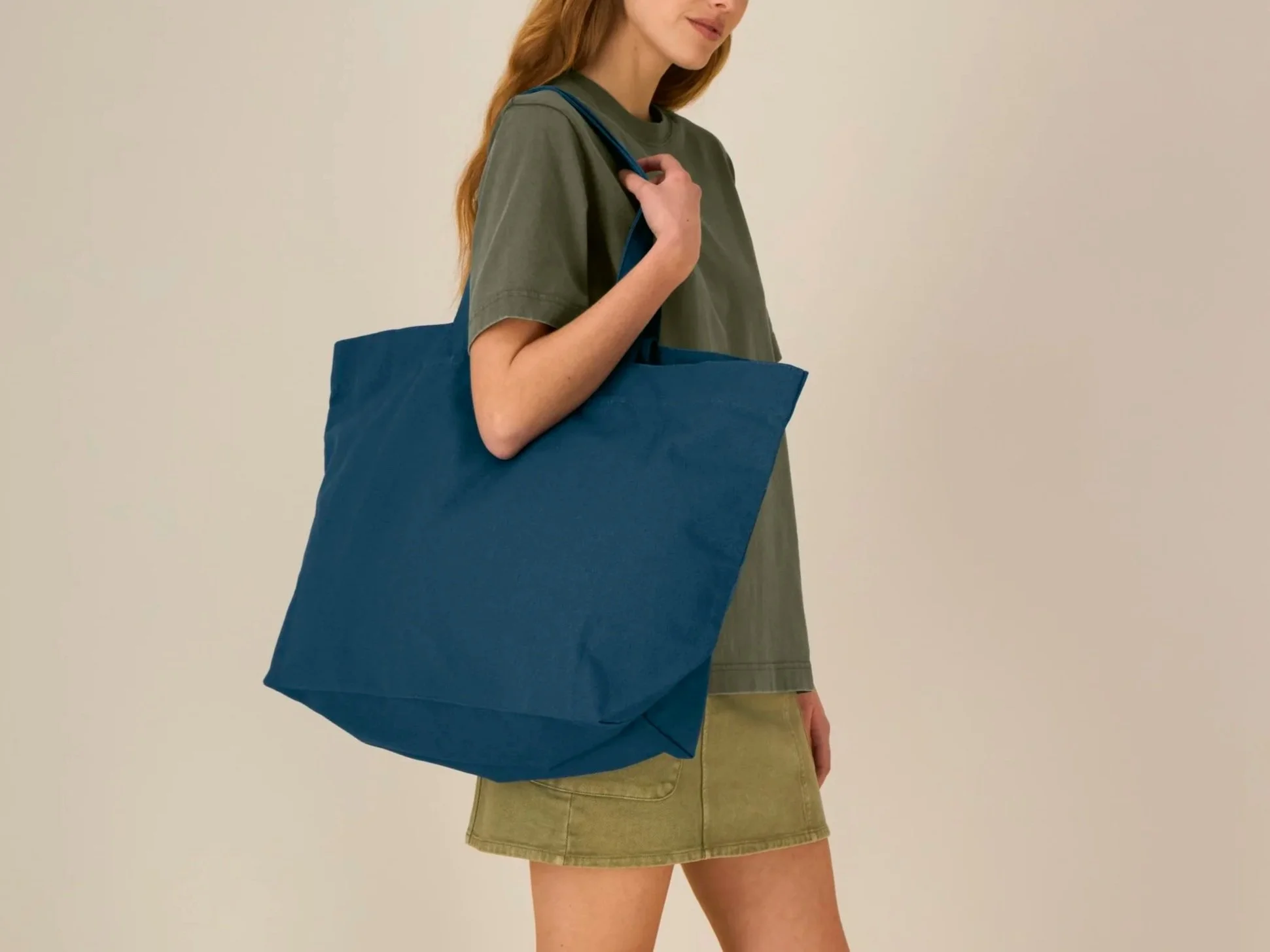 Woman with long red hair holding a large blue tote bag, wearing a loose gray-green T-shirt and a short khaki skirt, standing against a plain wall.