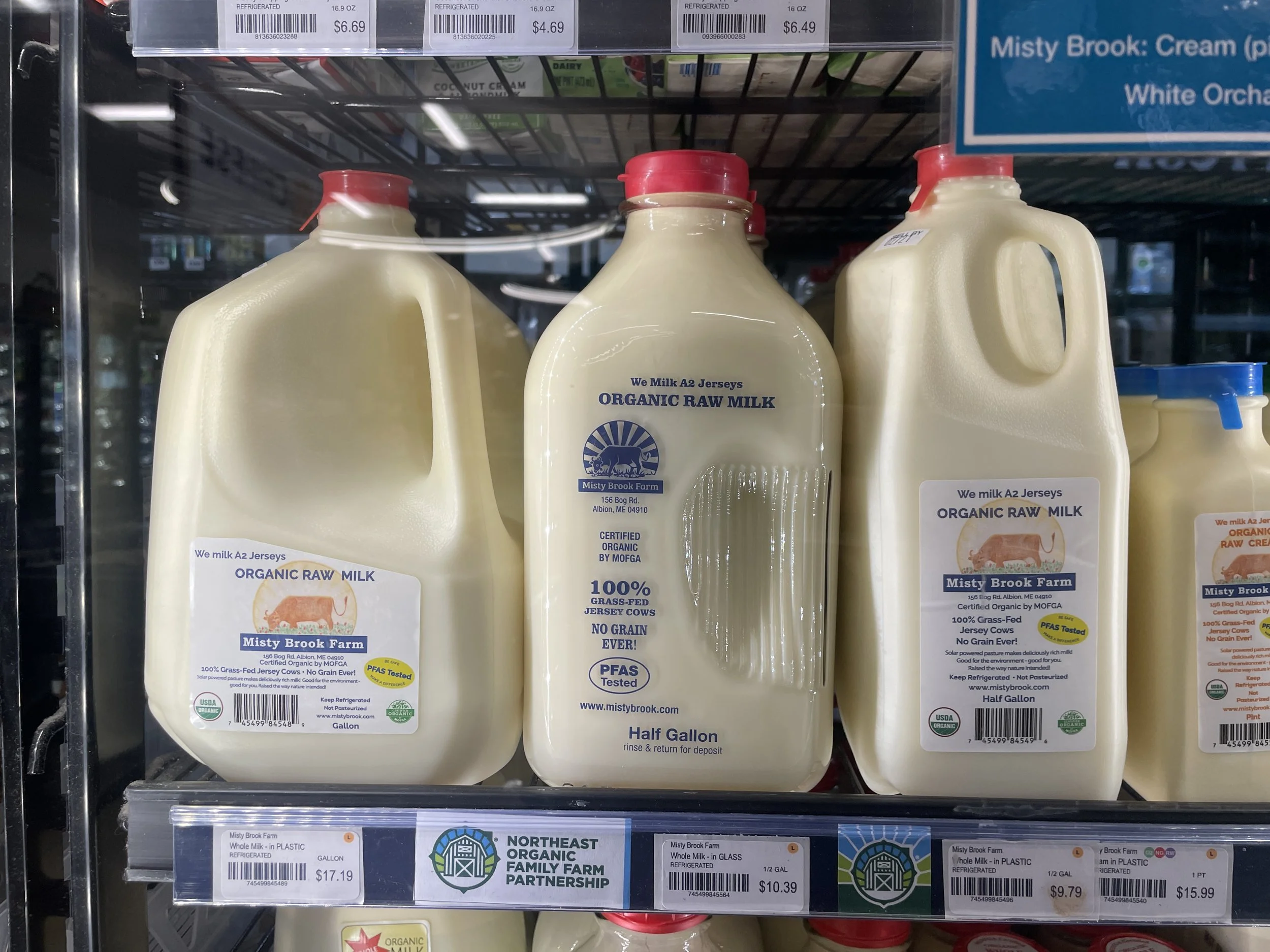 Three bottles of raw milk sit behind a glassed grocery store dairy aisle. They all have detailed labels, each of which include "raw milk."