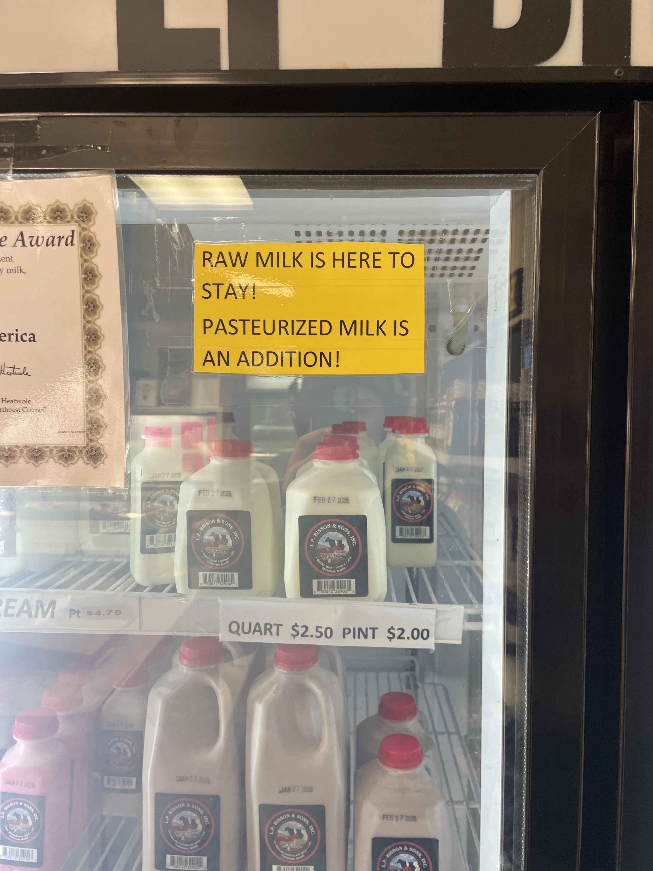 A glassed refrigerator stores pints of raw and pasteurized milk. A yellow sign affixed on the glass reads "RAW MILK IS HERE TO STAY! PASTEURIZED MILK IS AN ADDITION!"