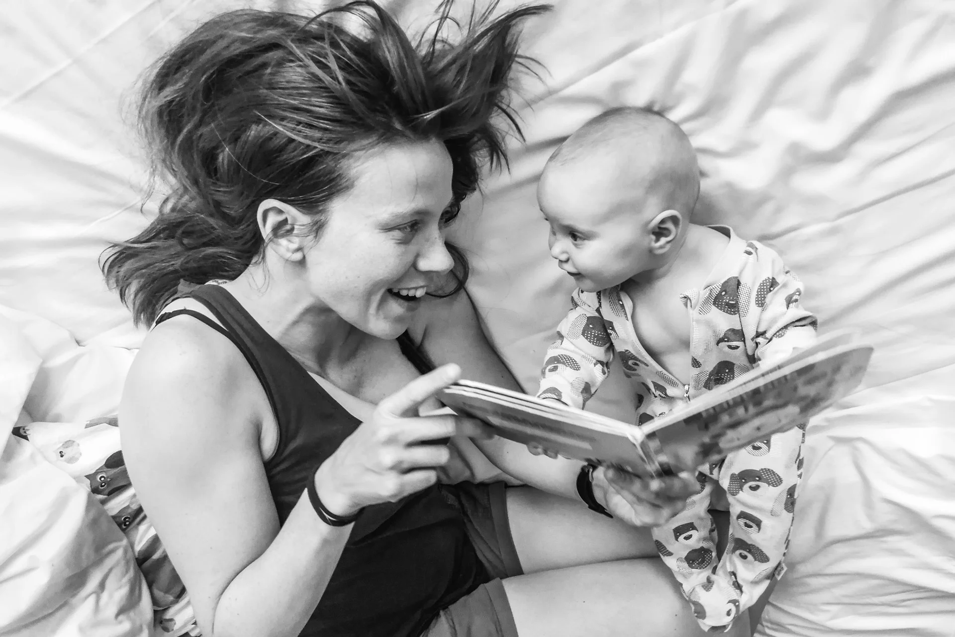 Mor og baby ligger på senga og leser bok. Ser på hverandre og smiler.