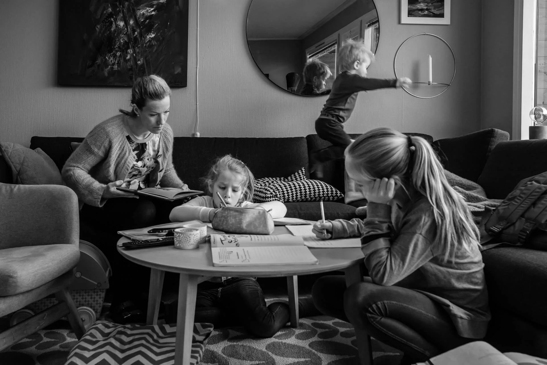 En barnefamilie er samlet rundt stuebordet. Mamma hjelper de to eldste jentene med leksene, mens lillebror hopper i sofaen.