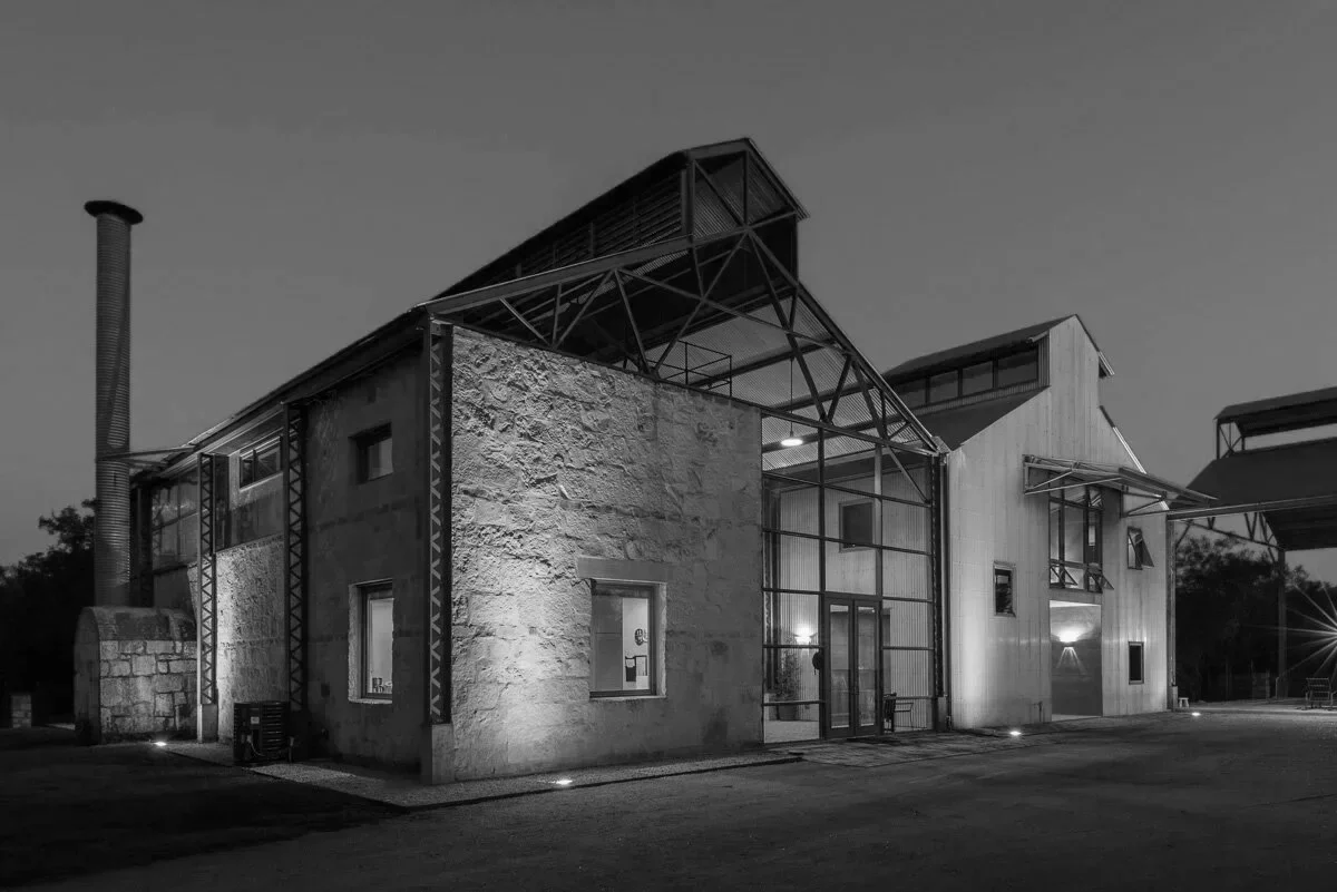 Black and white photo of a modern building with a mix of stone and metal siding, featuring large windows and a prominent chimney.