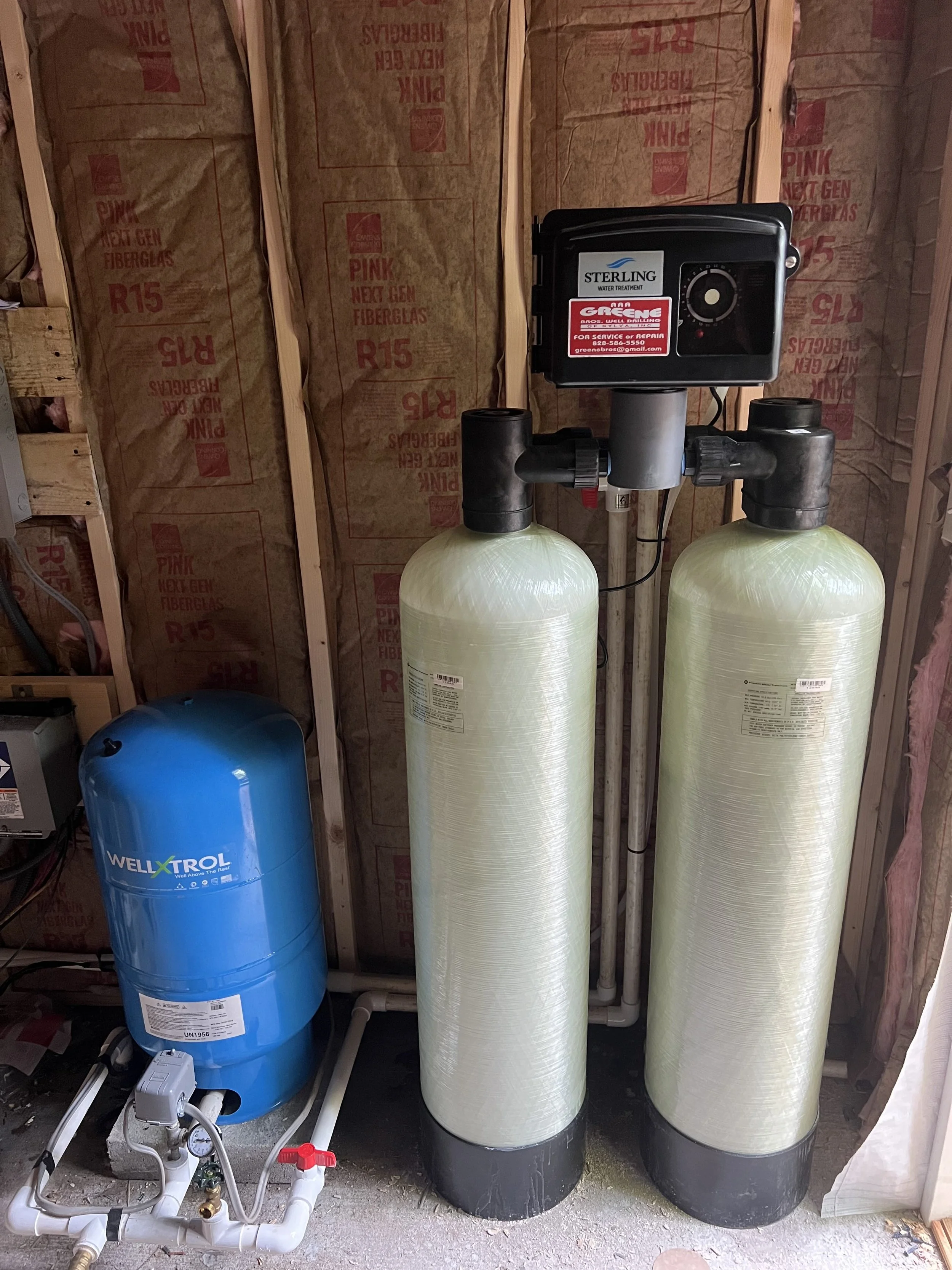 A water filtration system with two large beige tanks and a control valve on top, next to a blue WellXTrol pressure tank, all installed in a basement with unfinished walls.