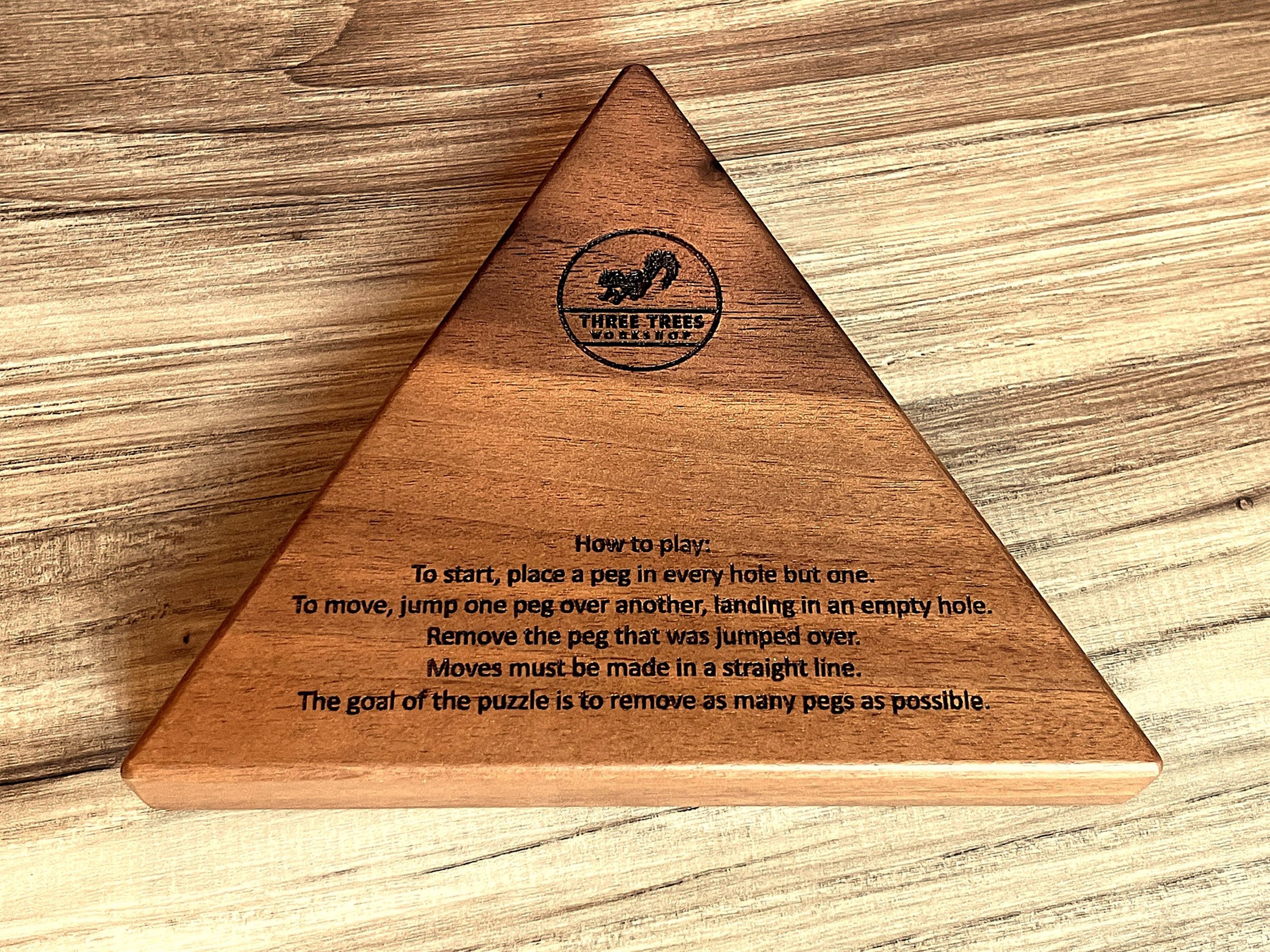 The back of a triangle solitaire board, with instructions and the Three Trees Workshop squirrel logo engraved in the wood.