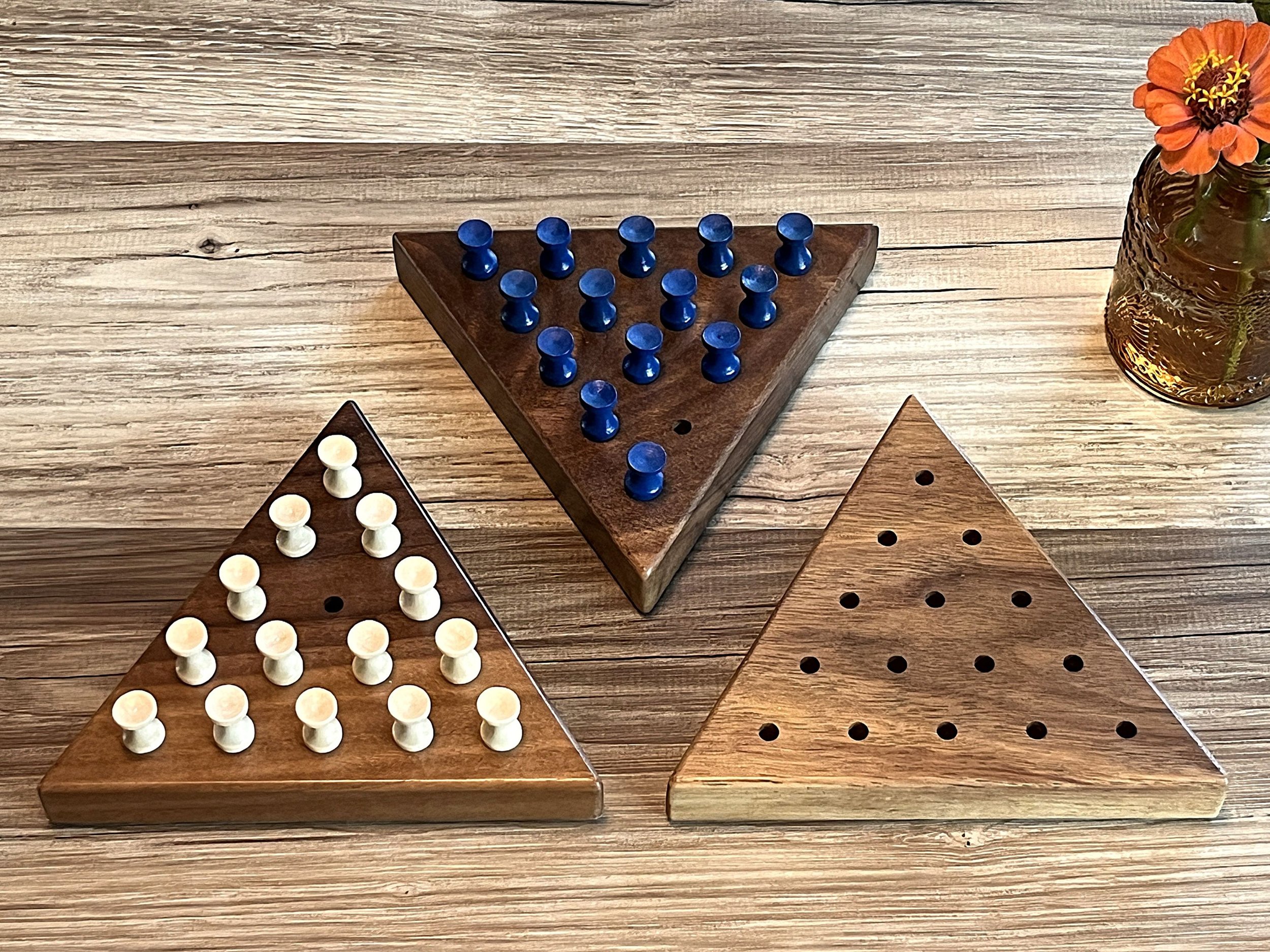 Three triangle peg solitaire puzzle boards side by side on a table. One has natural colored pegs in it, one has blue pegs, and the other has no pegs.