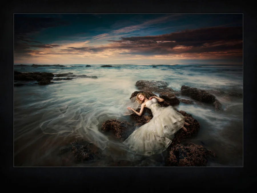 A Des Moines bride lays on a rock in the ocean and watches her veil wash back and forth in the tide. Destination wedding photographed in Oregon by Iowa Wedding Photographer of the Year McClanahan Studio. West coast wedding, dramatic sunrise portrait was selected for the 2014 PPA Loan Collection.