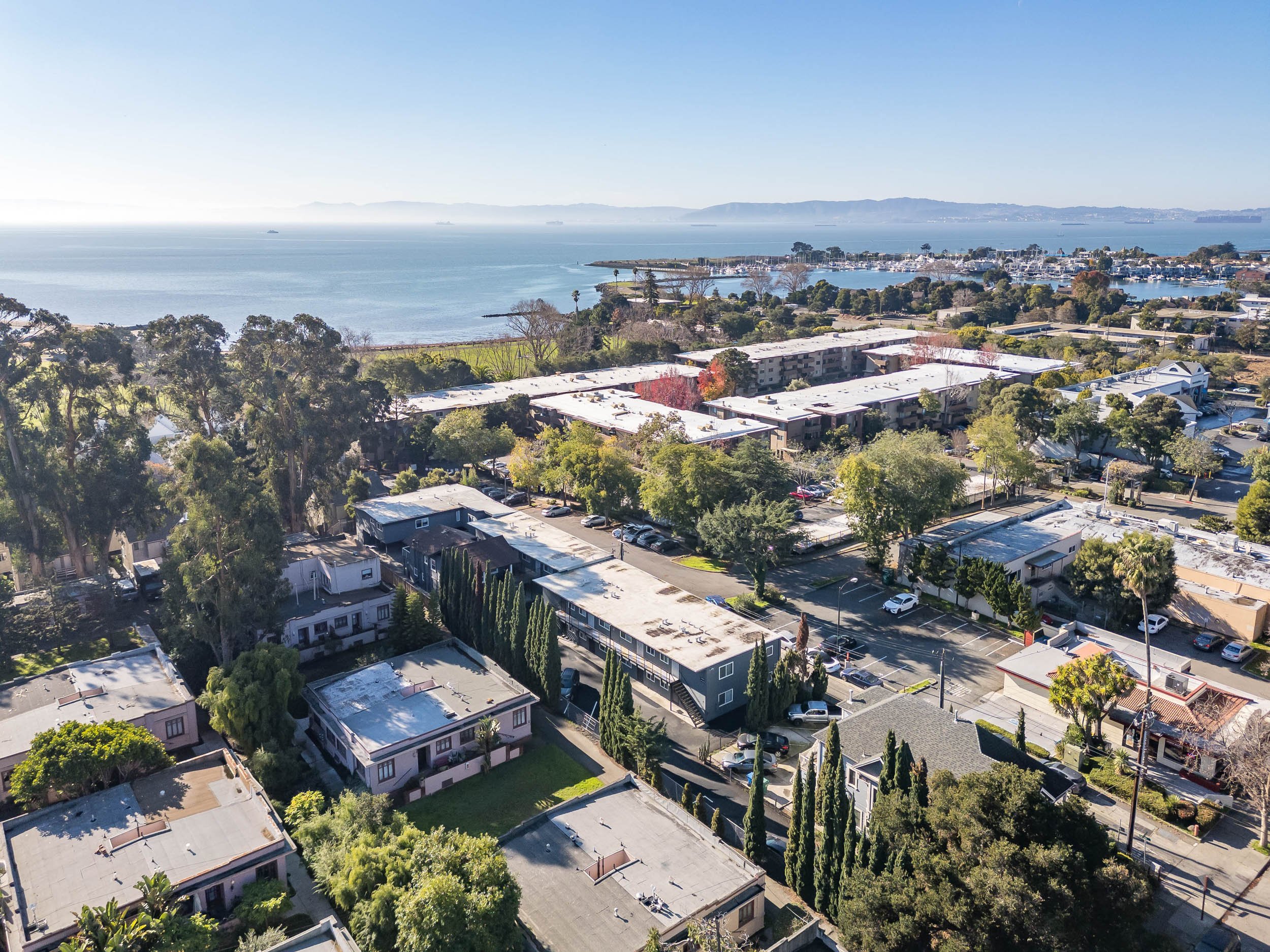 Aerial view of a coastal neighborhood with residential buildings, surrounded by trees and greenery. The ocean is visible in the background, along with a marina and distant hills under a clear blue sky.