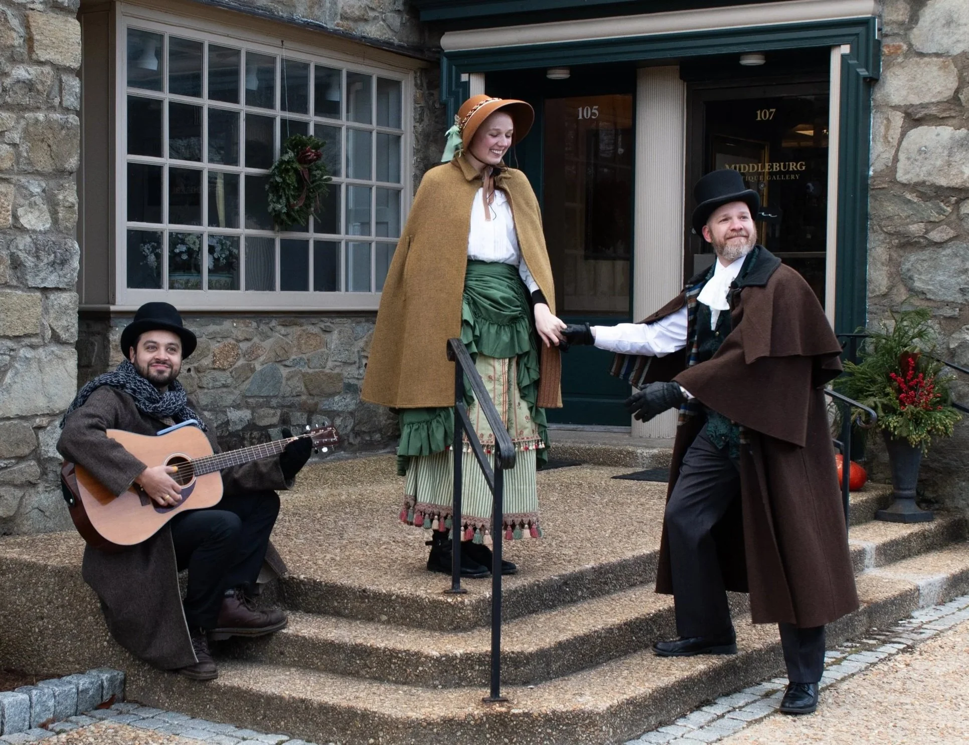 a couple in 1800s clothing smile at each other while a musical with a guitar  sits on the steps low them, also in 1800s clothing. they are all smiling