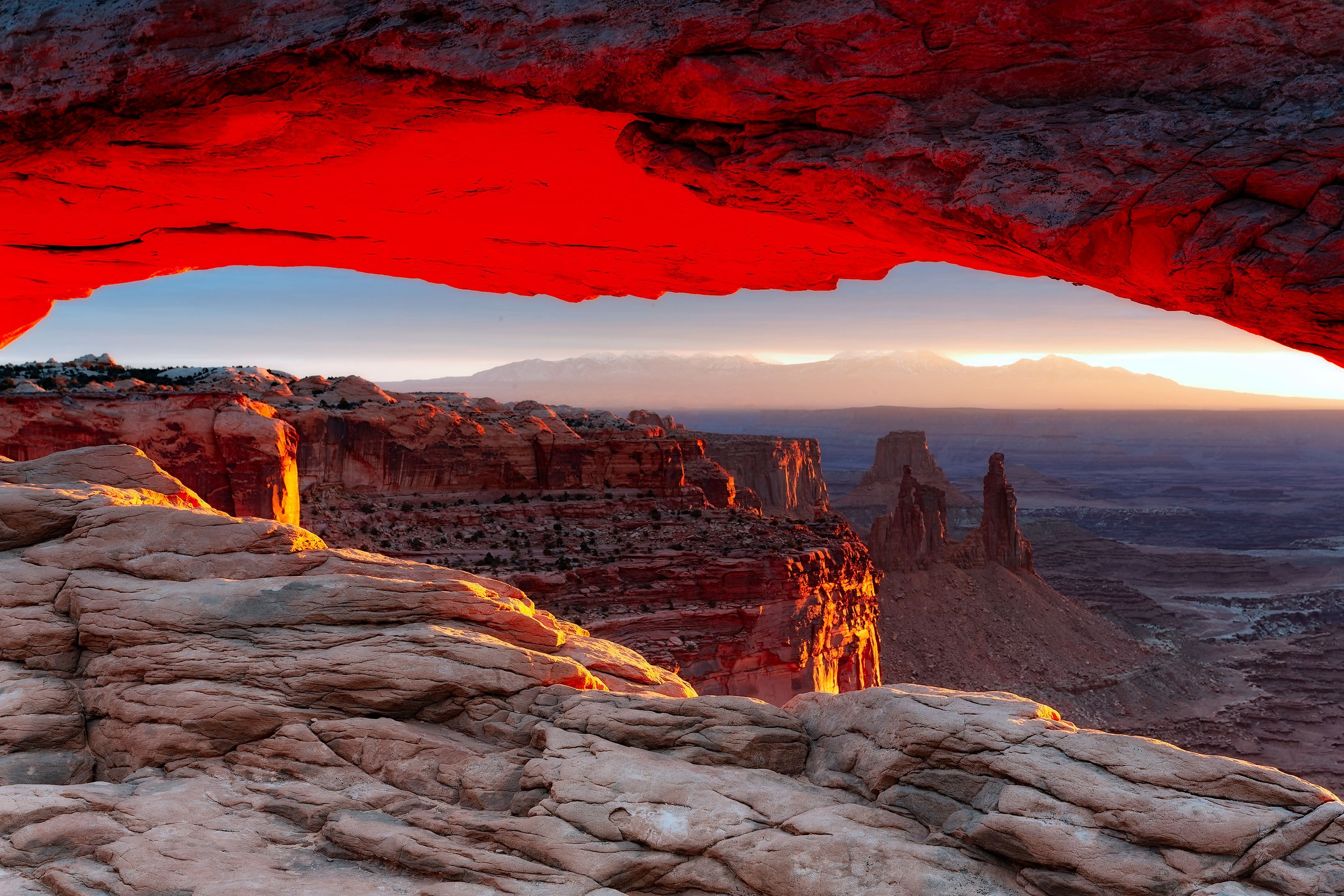 Sunrise light glowing under Mesa Arch in Canyonlands National Park, Utah, with desert cliffs and La Sal Mountains in the distance.