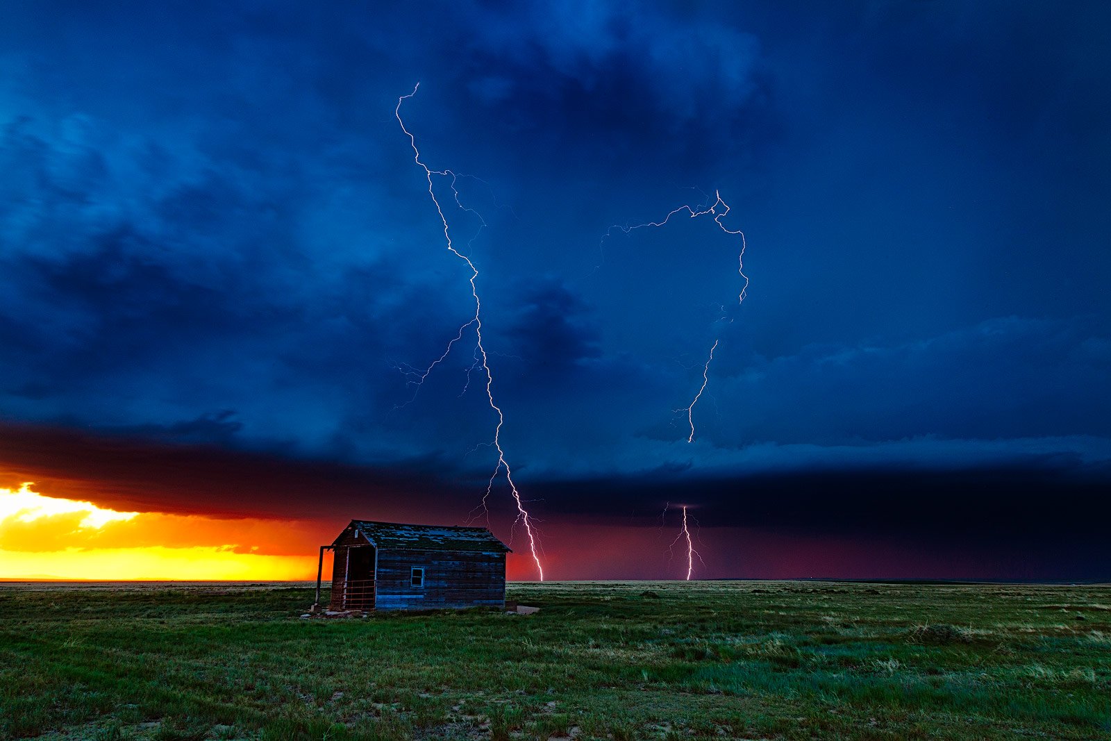 ightning bolts strike beneath a dramatic stormy sky on the Colorado plains, with a small weathered cabin in the foreground and a vibrant sunset glowing on the horizon.
