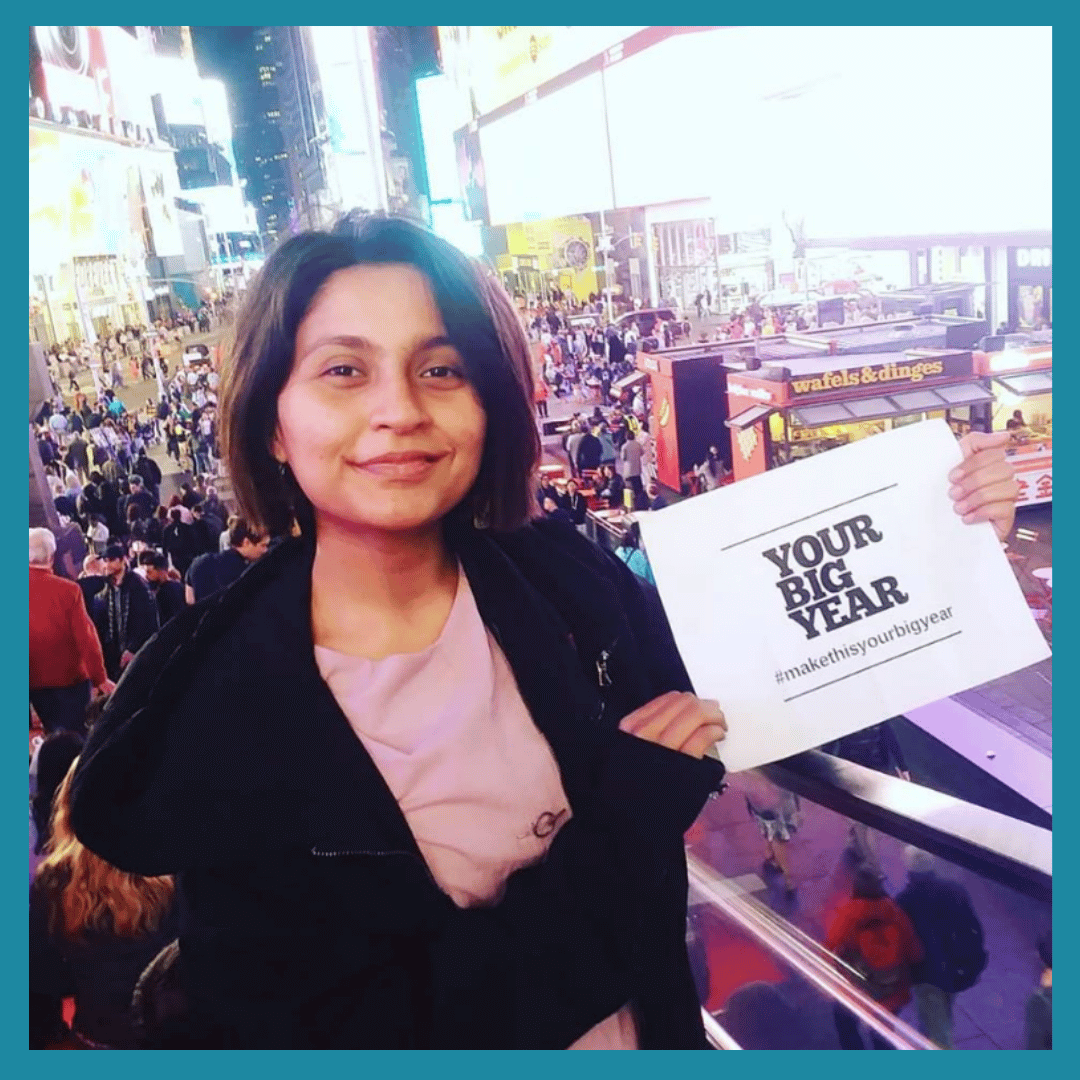 A woman smiling and holding a sign that says 'Your Big Year' with a hashtag #makethisyourbigyear, standing in a busy, brightly lit city area with crowds and tall buildings, possibly Times Square at night.