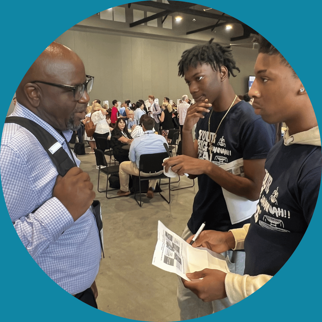 Three young men having a conversation at a busy indoor event or conference, with many people seated and standing in the background.