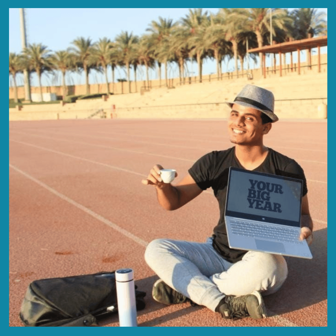 A man sitting cross-legged on a running track, holding a small cup in one hand and a laptop in the other, with a palm tree-lined background. The laptop screen displays the words 'YOUR BIG YEAR', and there is a water bottle and a bag nearby.
