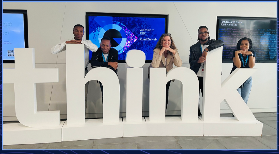 Five people are posing behind a large white sign that spells 'think' at a technology event, with digital screens in the background.