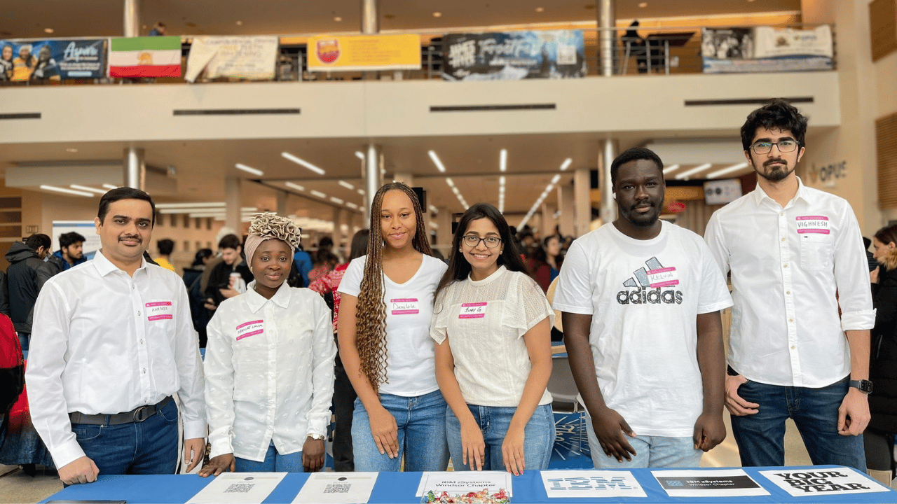 Group of six diverse people standing behind a table with signs and materials in a busy indoor event space.