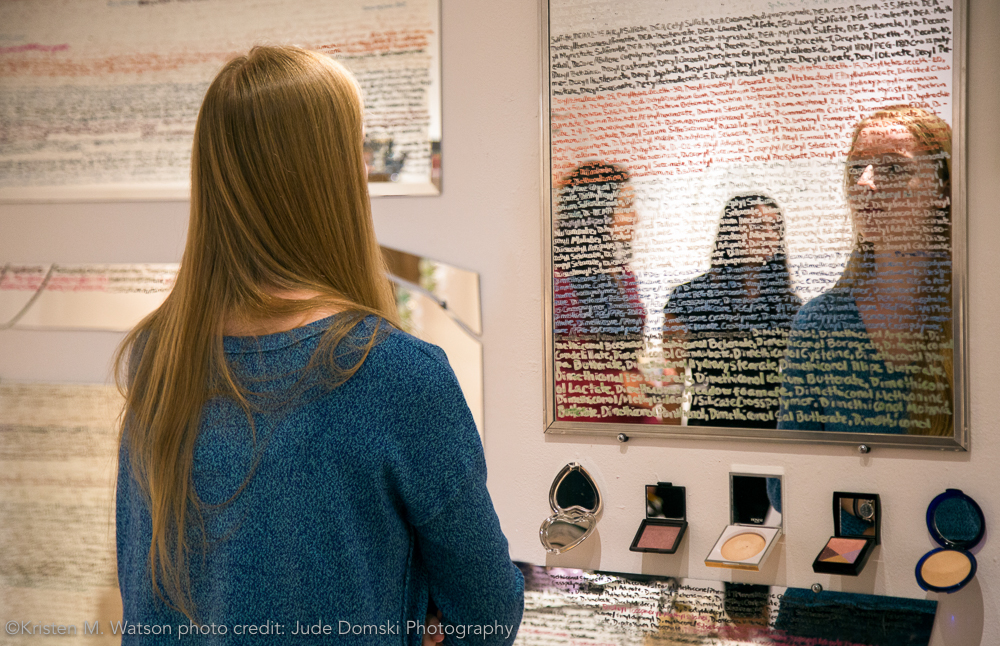 Visitor interacting with rəˈflekt , kristen m. watson, 2016, cosmetics ingredients list written using make up on mirror, overall dimensions approximately 60" x 240". written alphabetically by mirror from left to right.