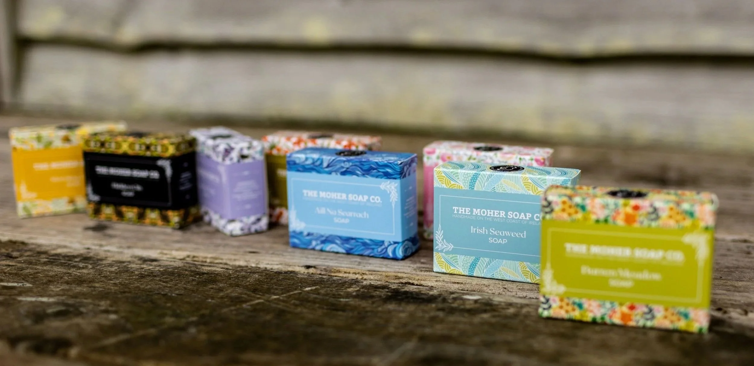 Small colorful soap bars labeled 'The Moher Soap Co.', including 'Irish Seaweed Soap', arranged in a row on a weathered wooden surface.